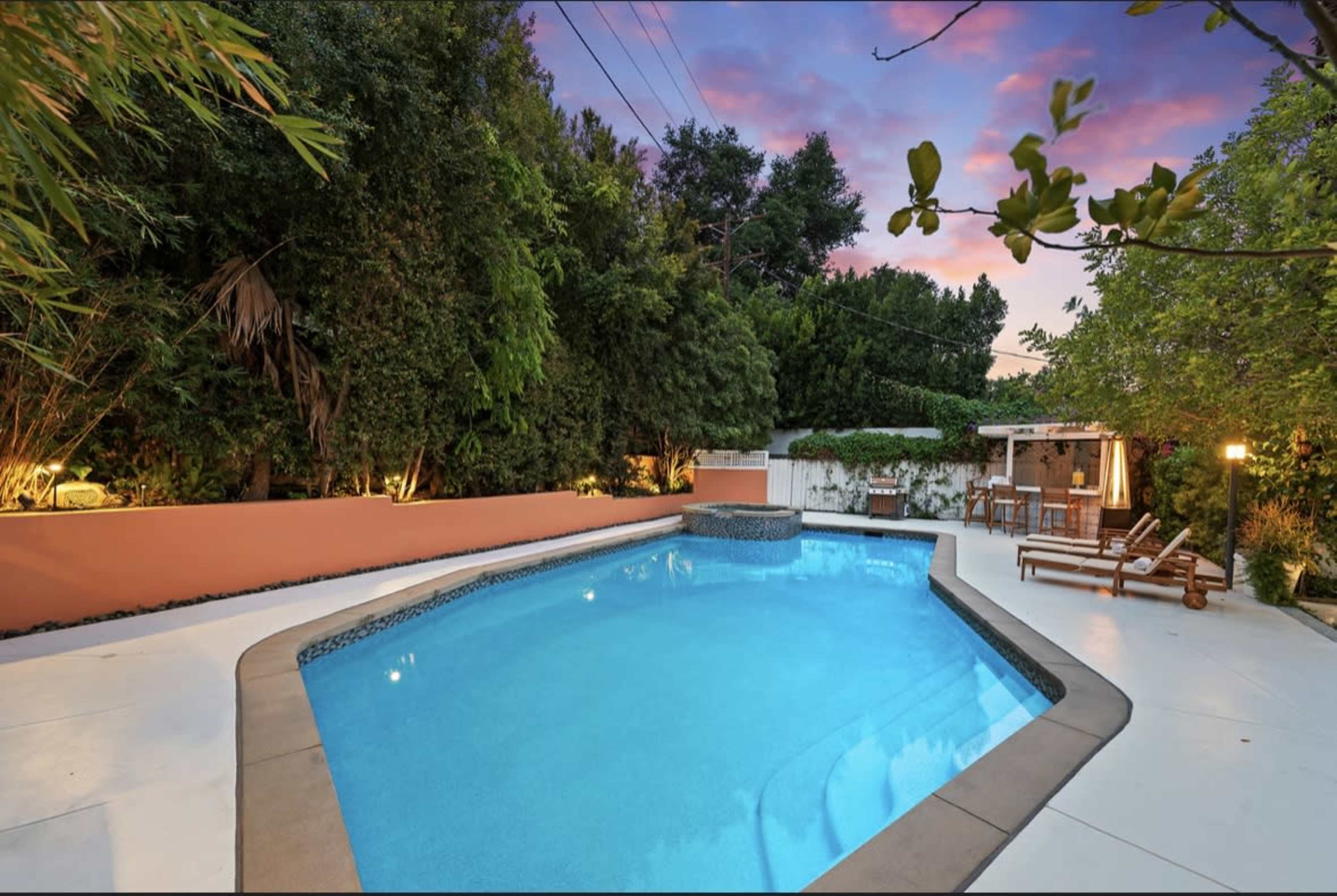 A backyard pool with a spa feature, surrounded by greenery and outdoor seating areas under a colorful sunset sky.