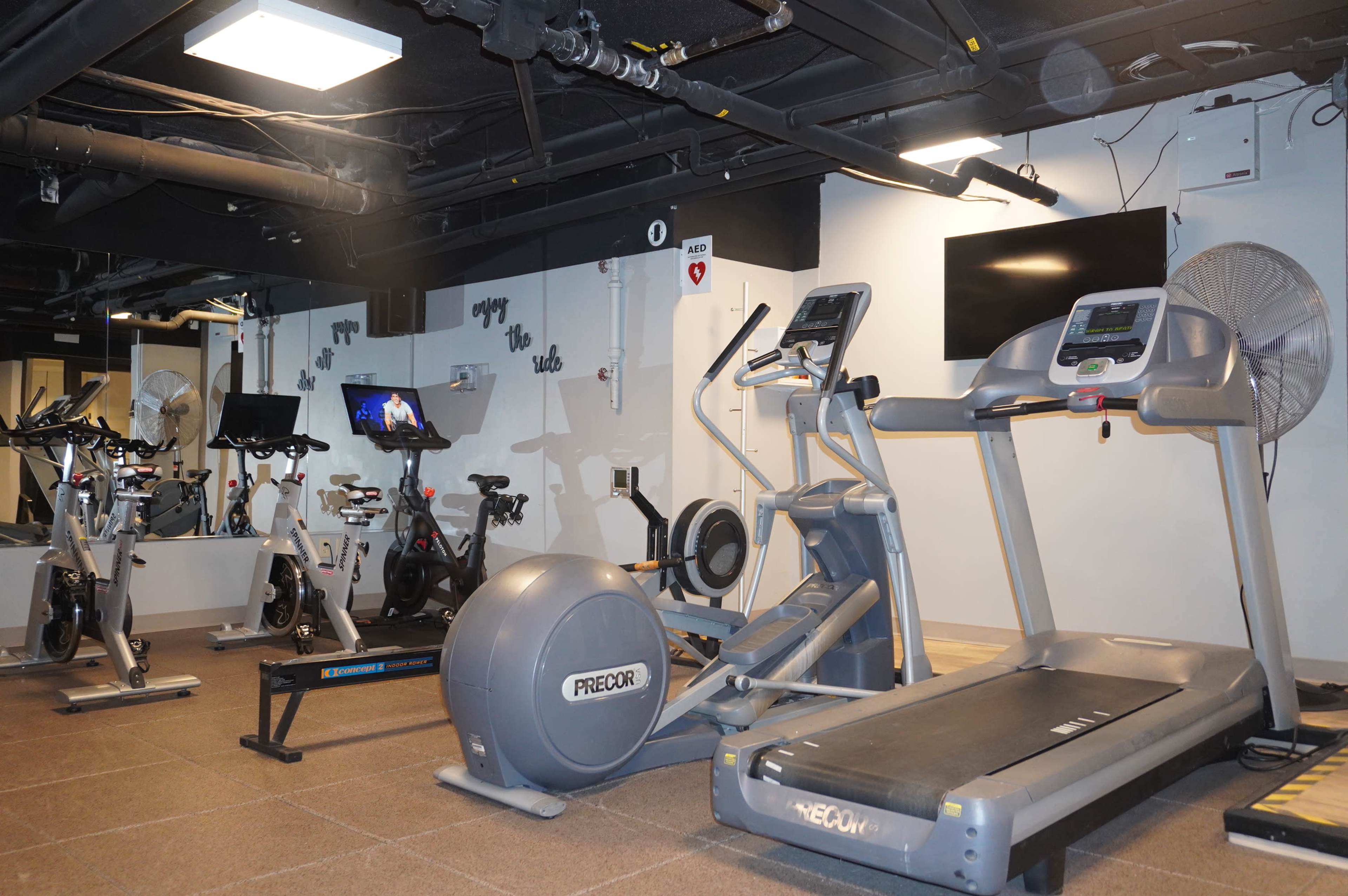 The image shows a modern gym area equipped with cardio machines, including treadmills, ellipticals, and stationary bikes, under a ceiling with exposed beams and ventilation.