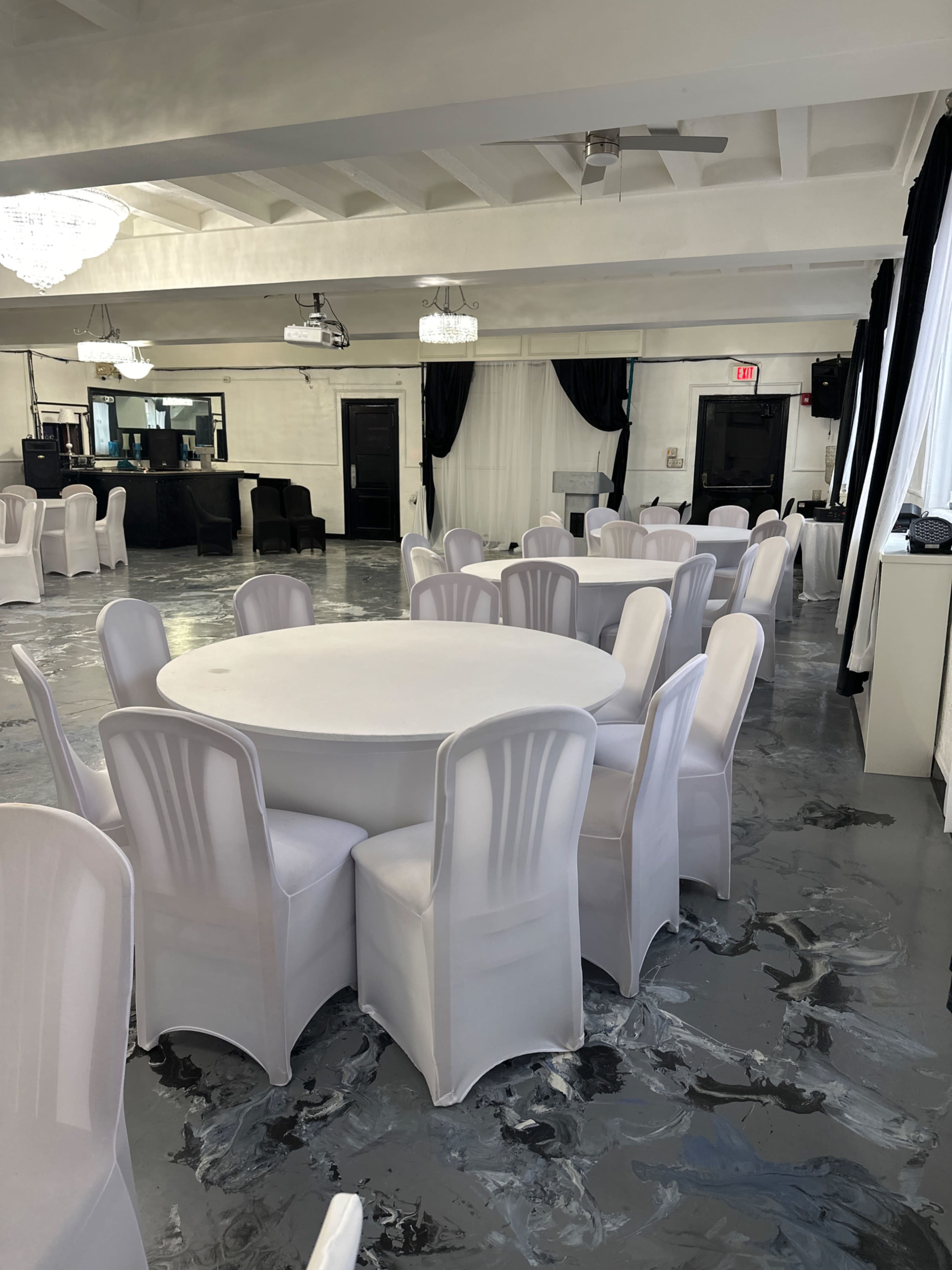 A spacious event room features round tables with white tablecloths and chairs, accented by black and white décor.