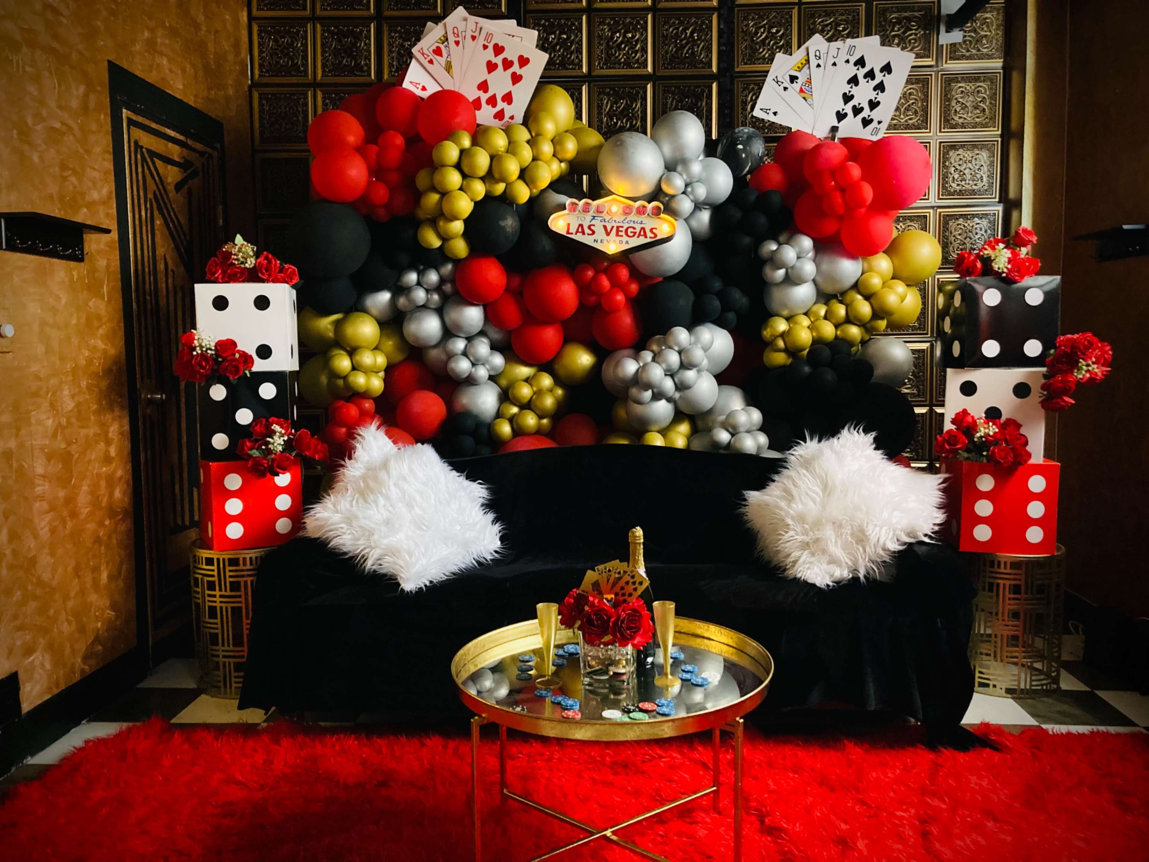 The image features a vibrant party setup with a black sofa, fluffy white pillows, a round golden coffee table, and a backdrop of colorful balloons arranged in a casino theme, accompanied by decorative elements like dice and card motifs.