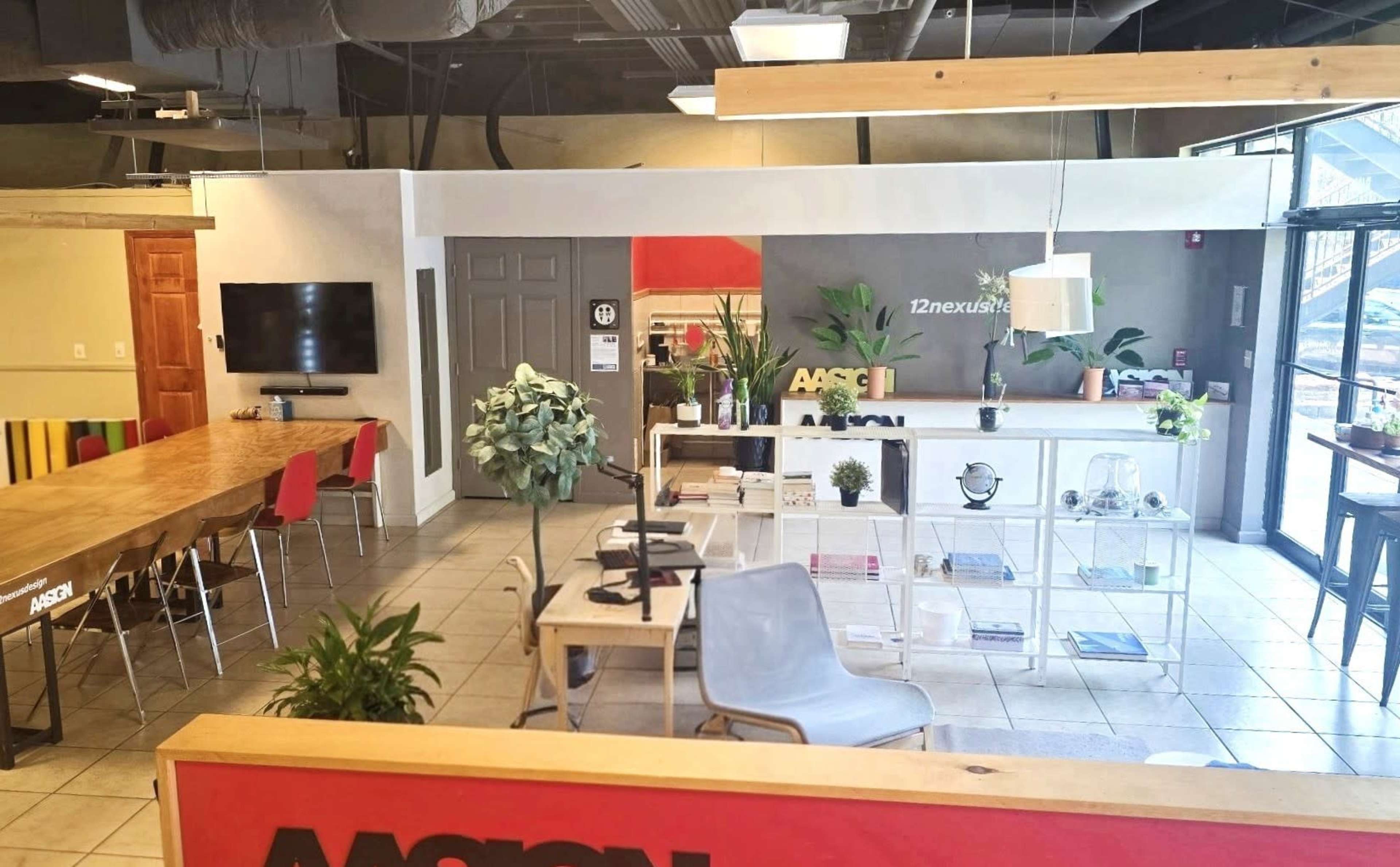 The image shows a modern office space with a large communal table, a wall-mounted TV, green plants, and various workstations, all arranged in a bright, open layout.