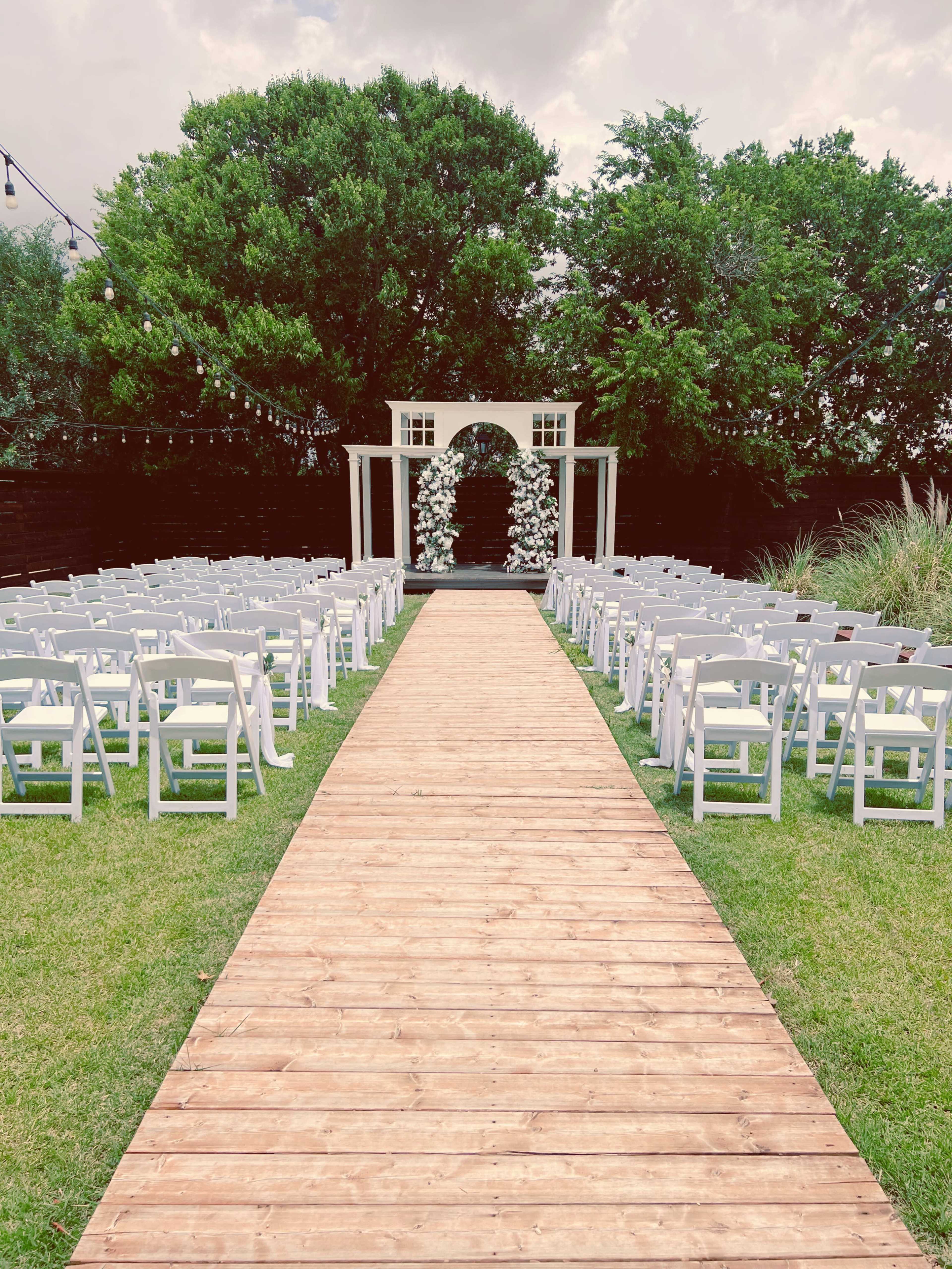 The image shows a wedding ceremony setup with white chairs arranged in rows on either side of a wooden walkway leading to a floral arch.