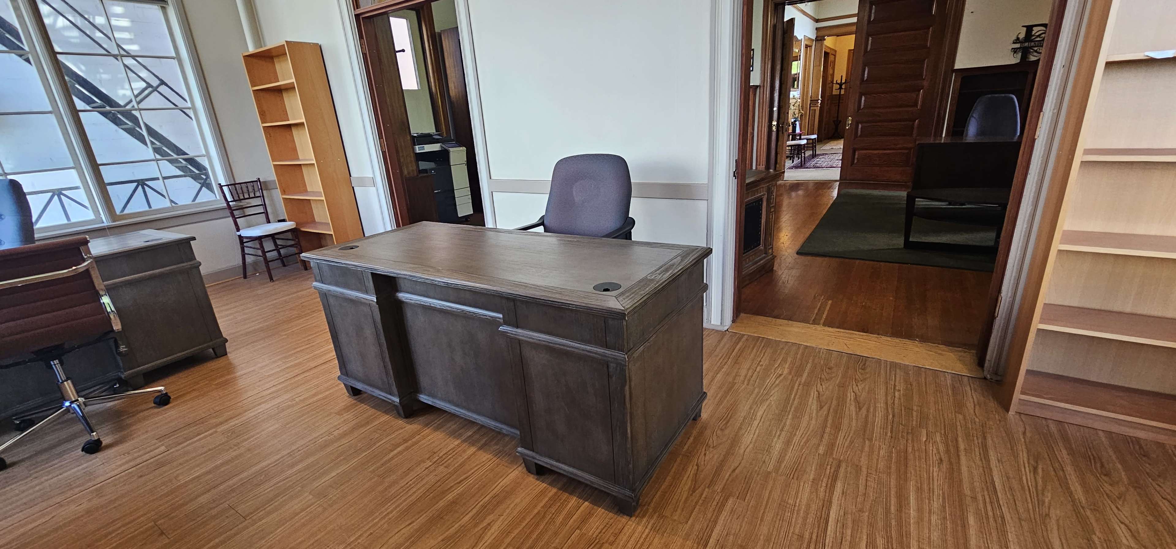 A wooden desk is positioned in an office space with large windows and a chair behind it, leading to an adjoining room.