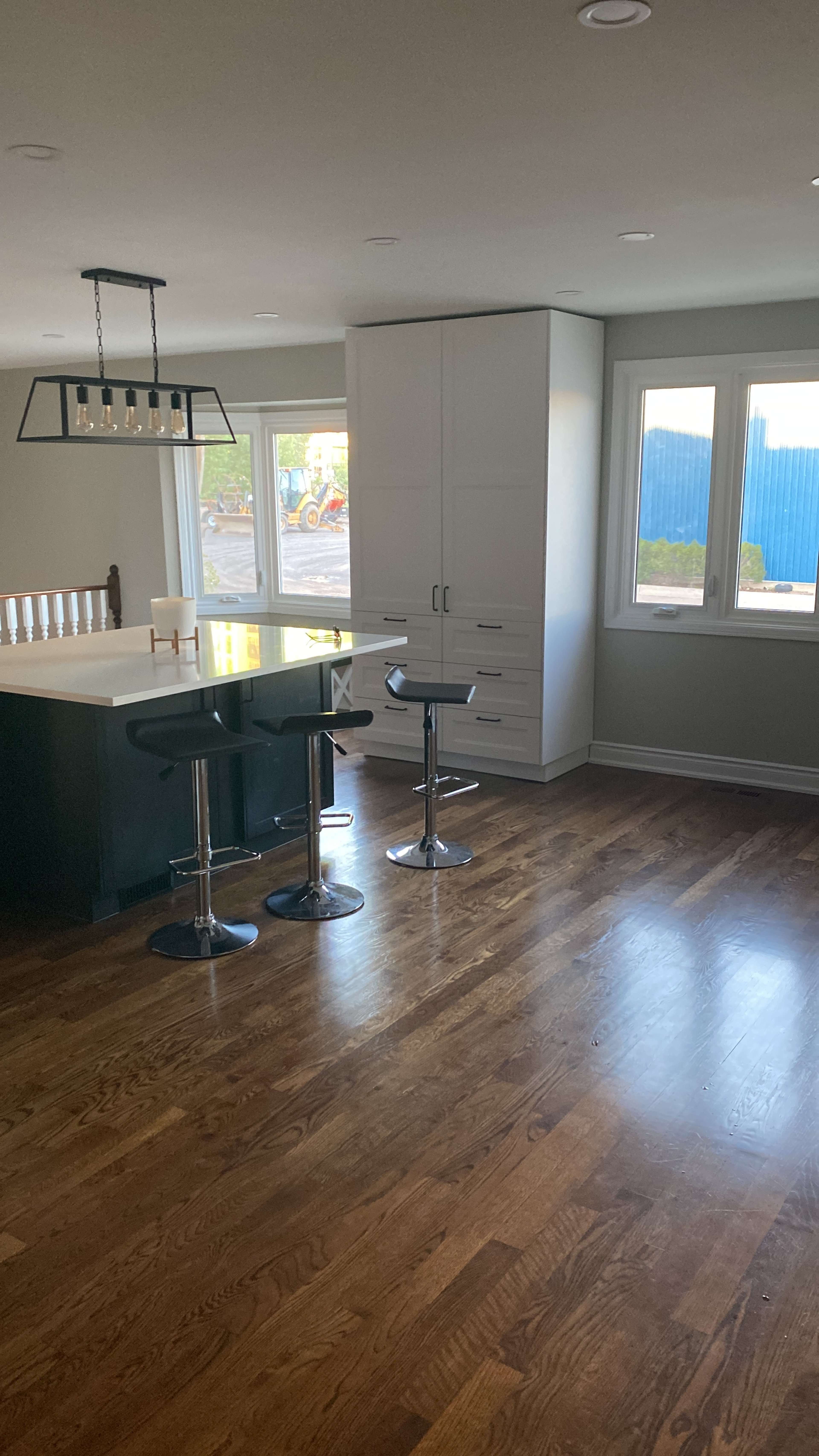 The image shows a modern kitchen area featuring a bar counter with two stools, large windows, and hardwood flooring.