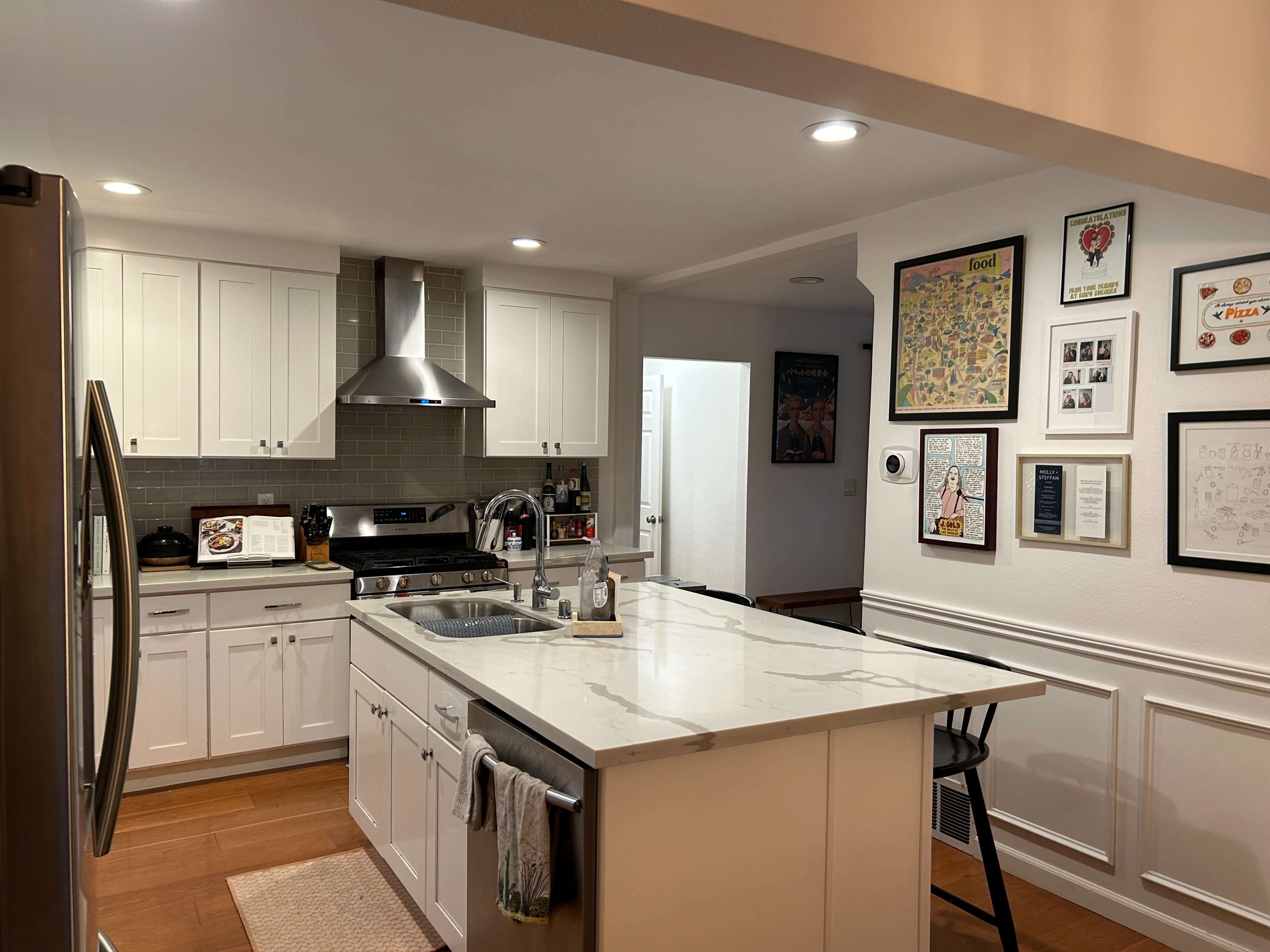 A modern kitchen with white cabinetry, a marble countertop, stainless steel appliances, and a wall adorned with framed artwork.