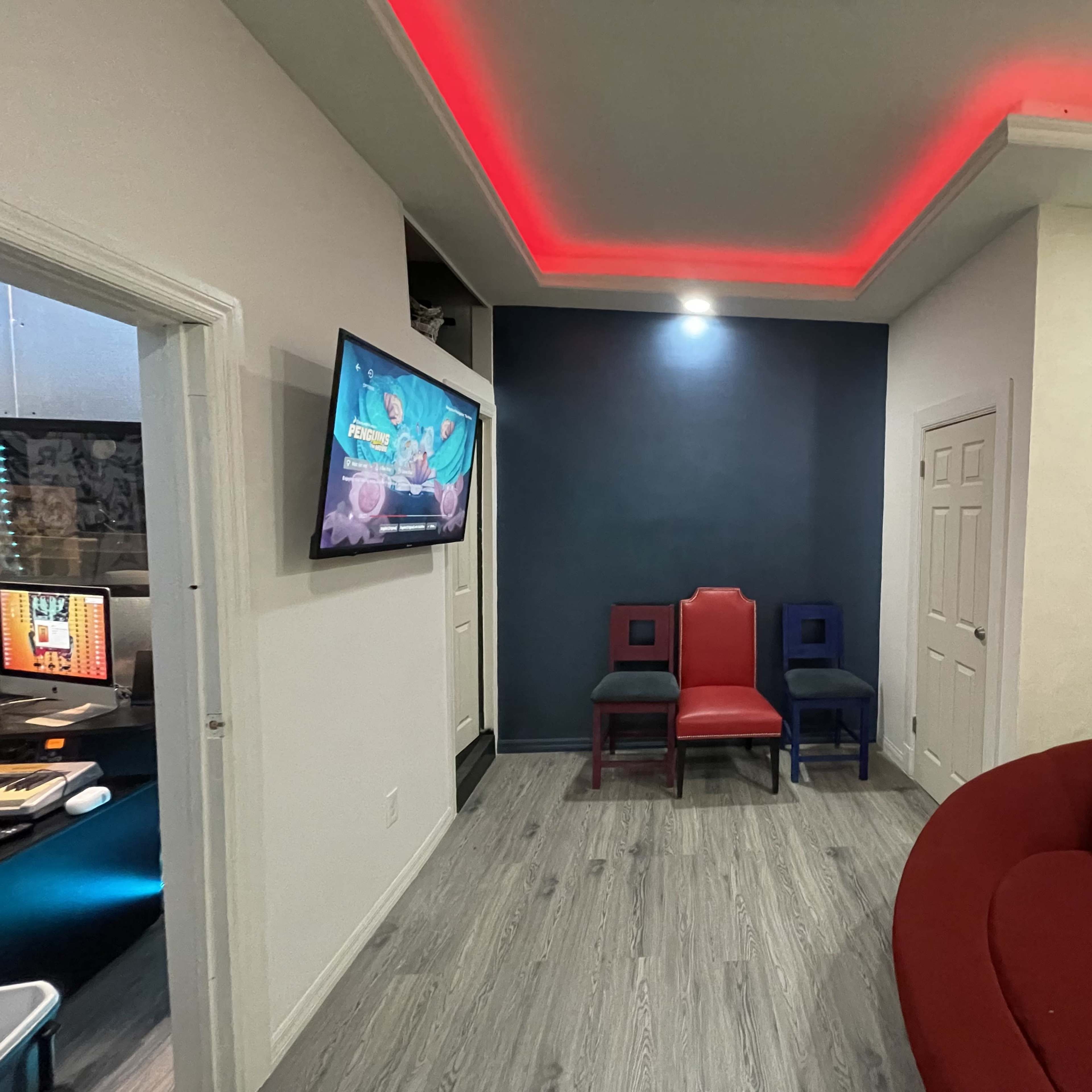 A hallway with a television mounted on the wall, two colored chairs (red and blue) beside it, and ambient red lighting along the ceiling.
