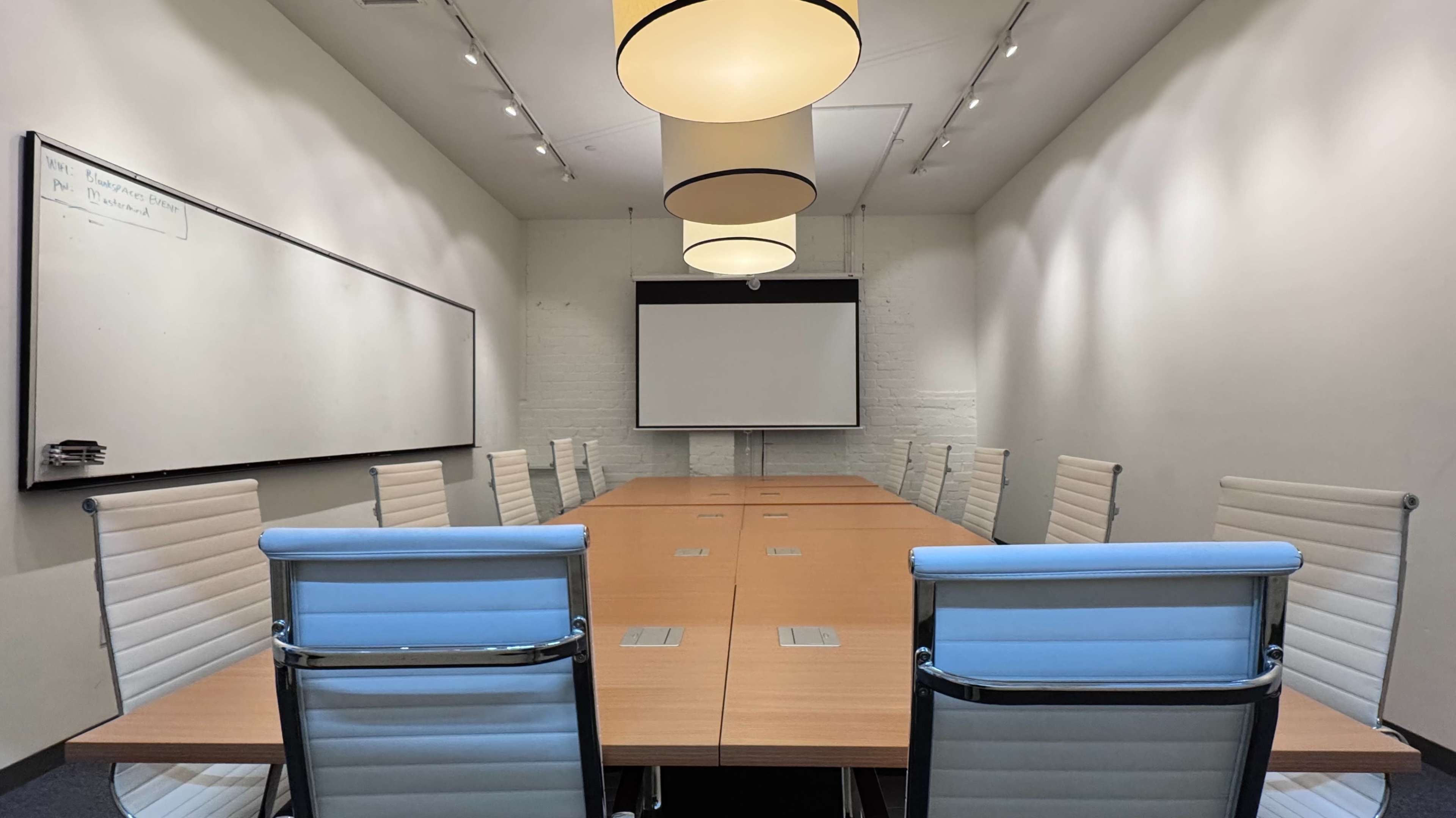 A long wooden conference table is surrounded by white chairs in a well-lit meeting room with a projector screen on one wall.