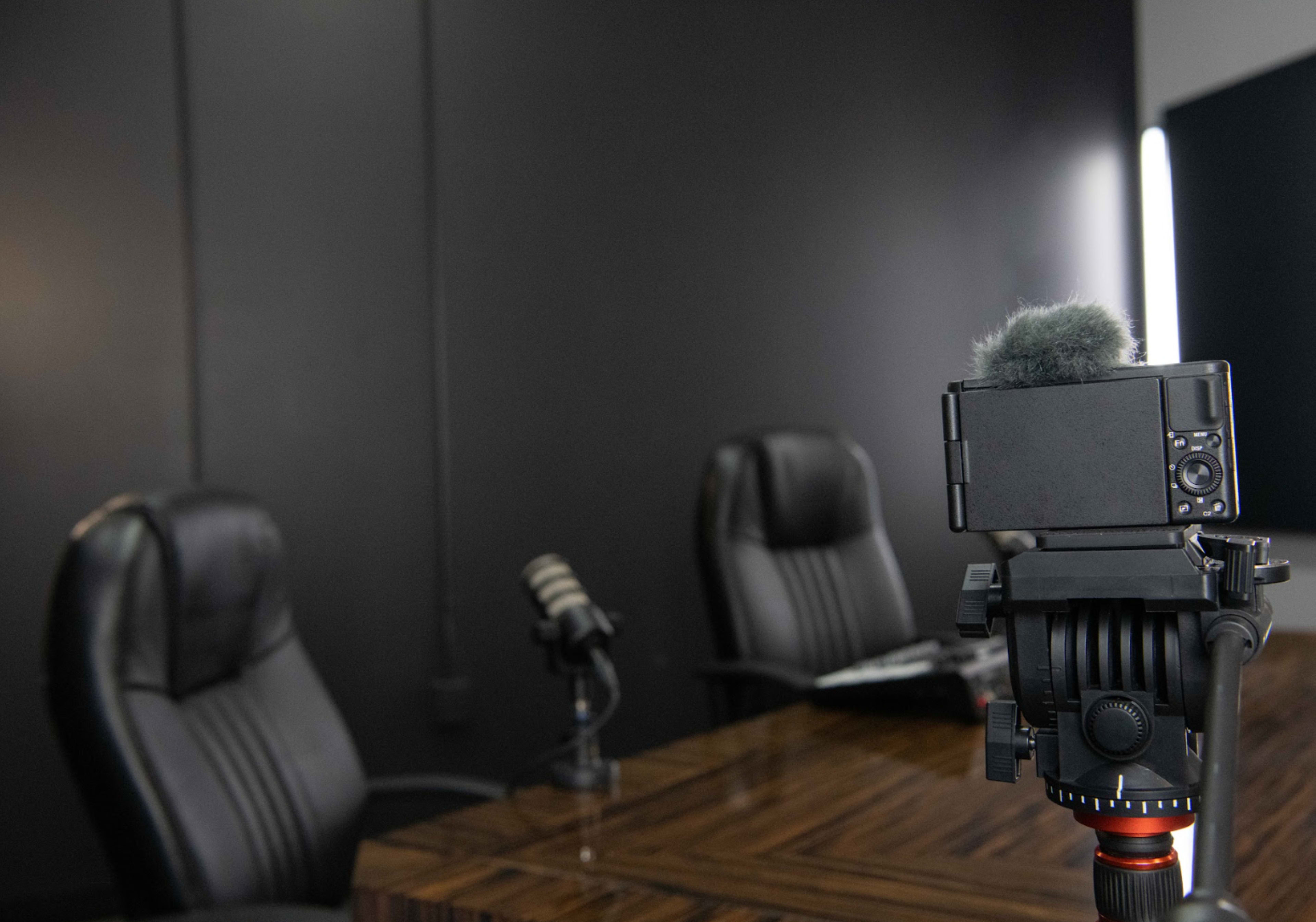 A camera with a microphone is positioned on a tripod facing two empty black leather chairs and a wooden table in a dimly lit room.