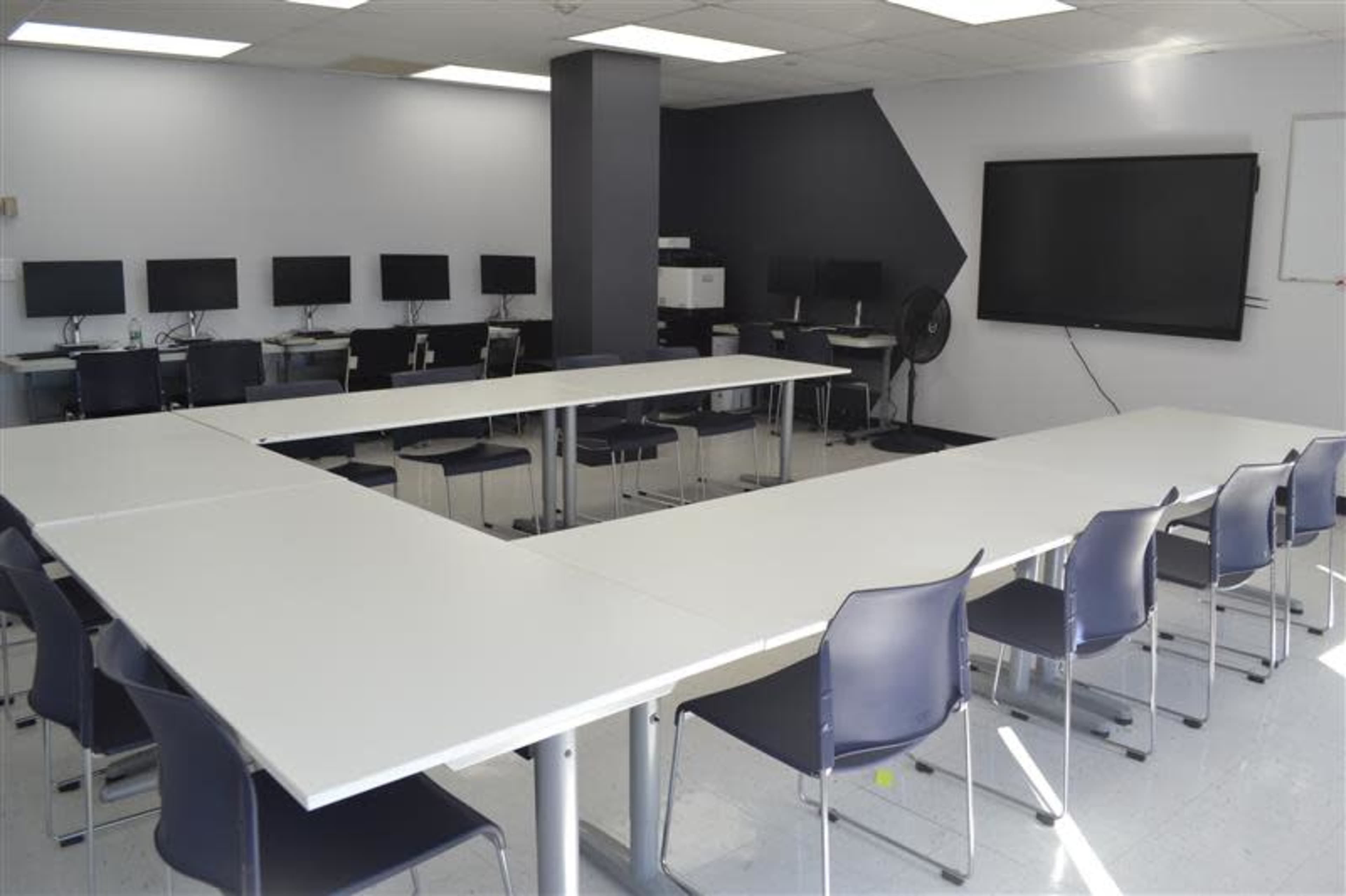 The image shows a classroom with several tables arranged in an "U" shape, surrounded by chairs, and equipped with computers against one wall.