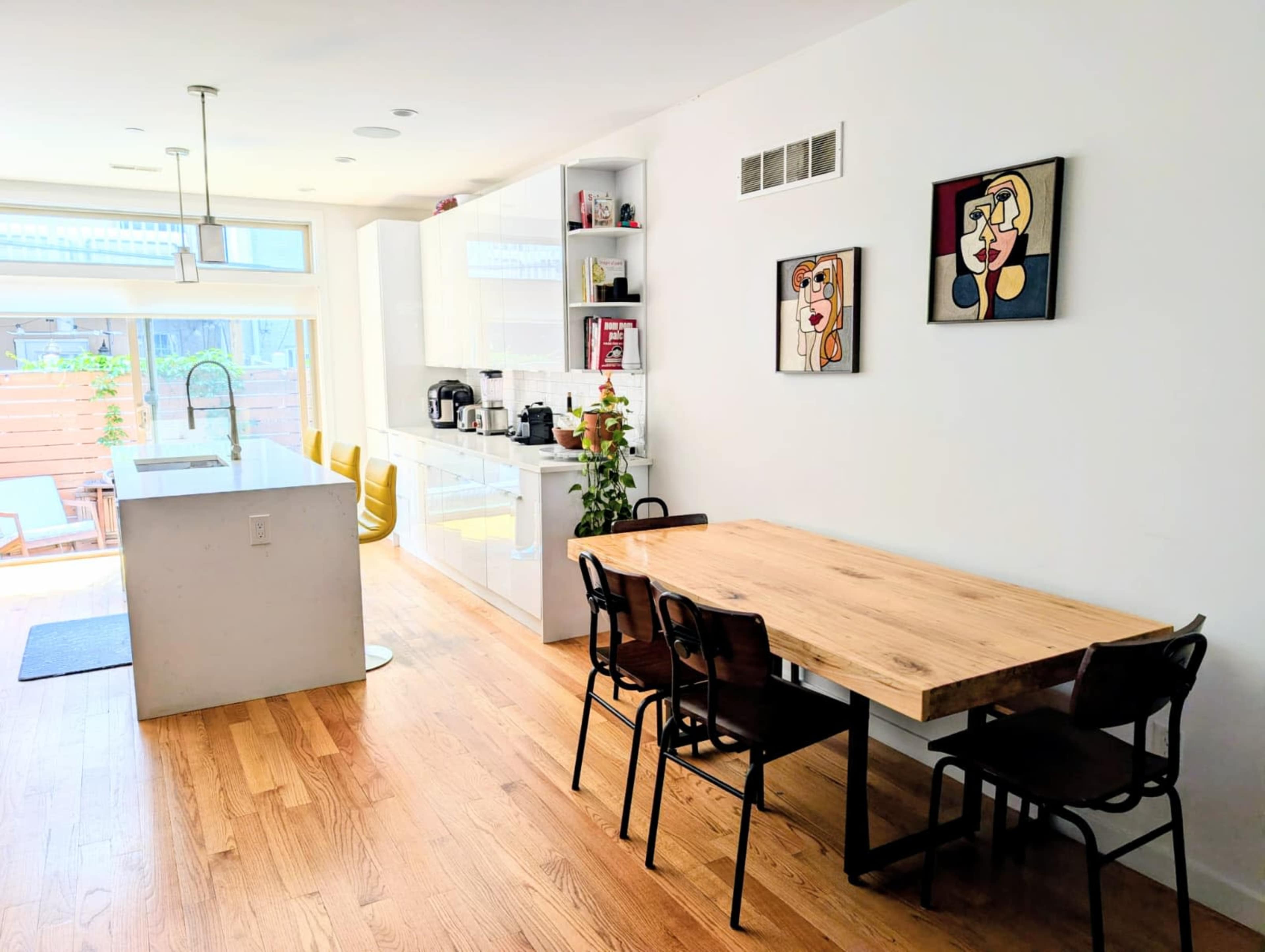 The image shows a modern kitchen and dining area with a wooden table and four chairs, complemented by minimalist decor and large windows allowing natural light.