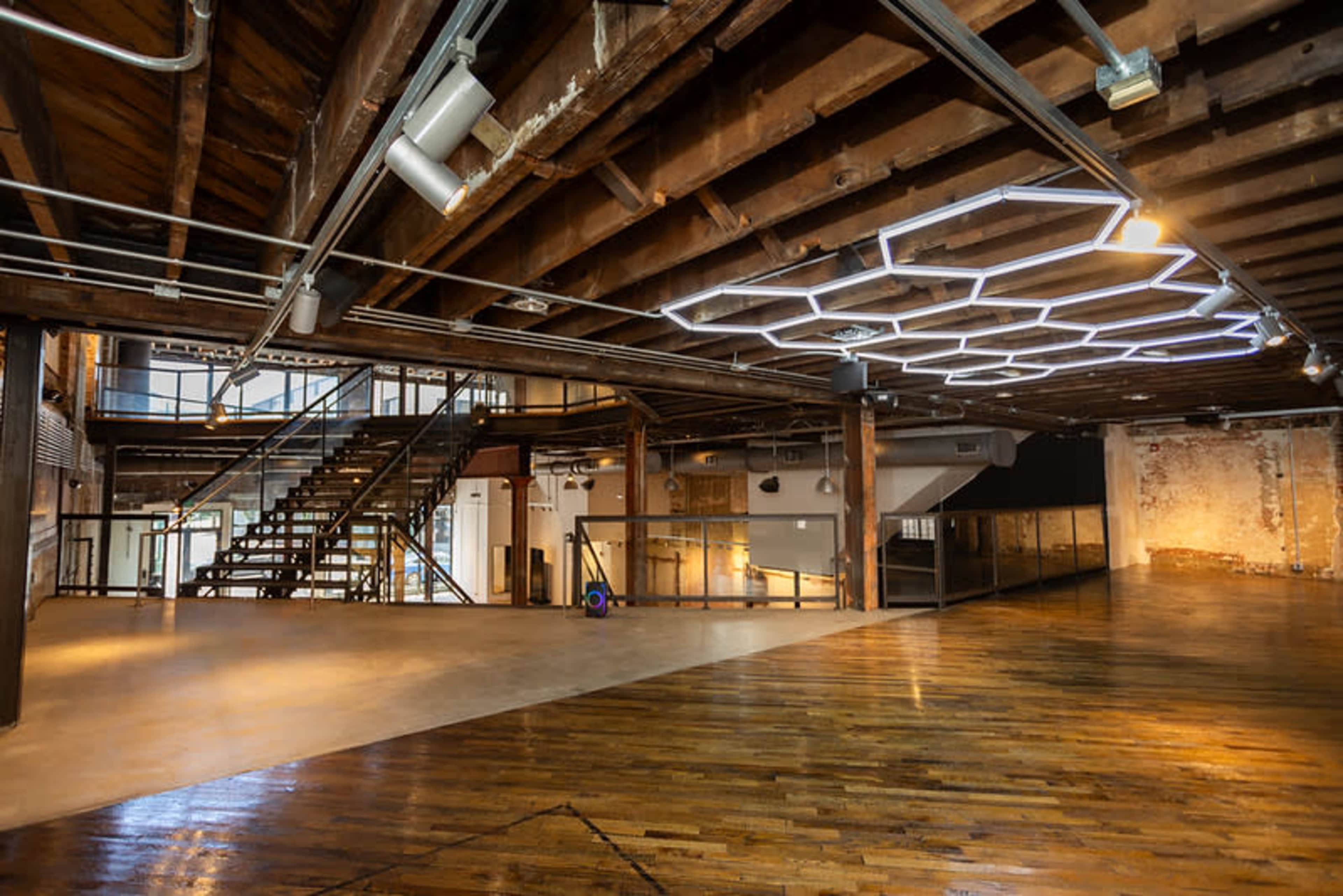 The image shows a spacious interior of a modern, industrial-style building with exposed wooden beams, a staircase, and a unique hexagonal light fixture on the ceiling.
