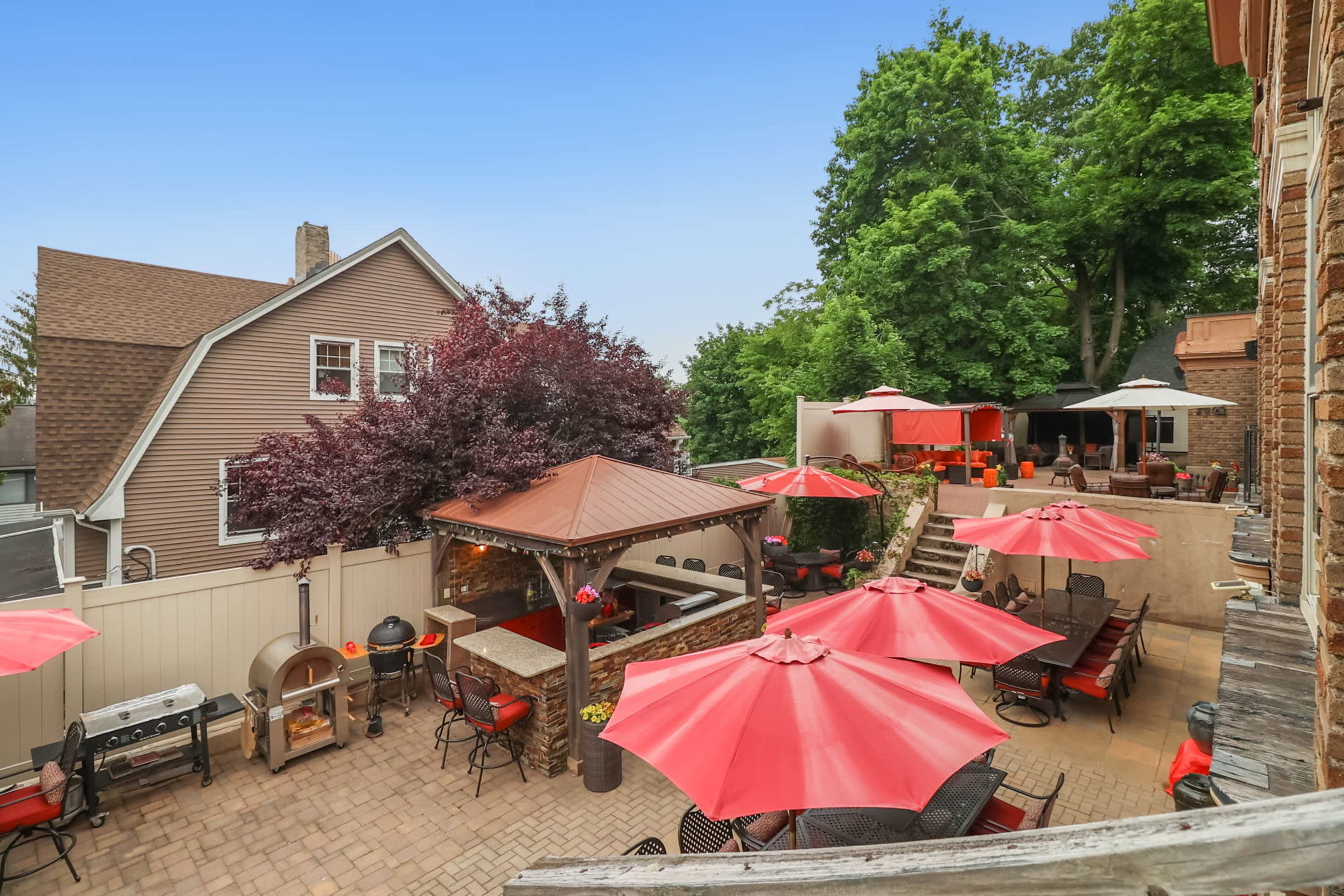 A patio area with red umbrellas, a stone bar, and a barbecue grill, surrounded by a well-maintained backyard and neighboring houses.