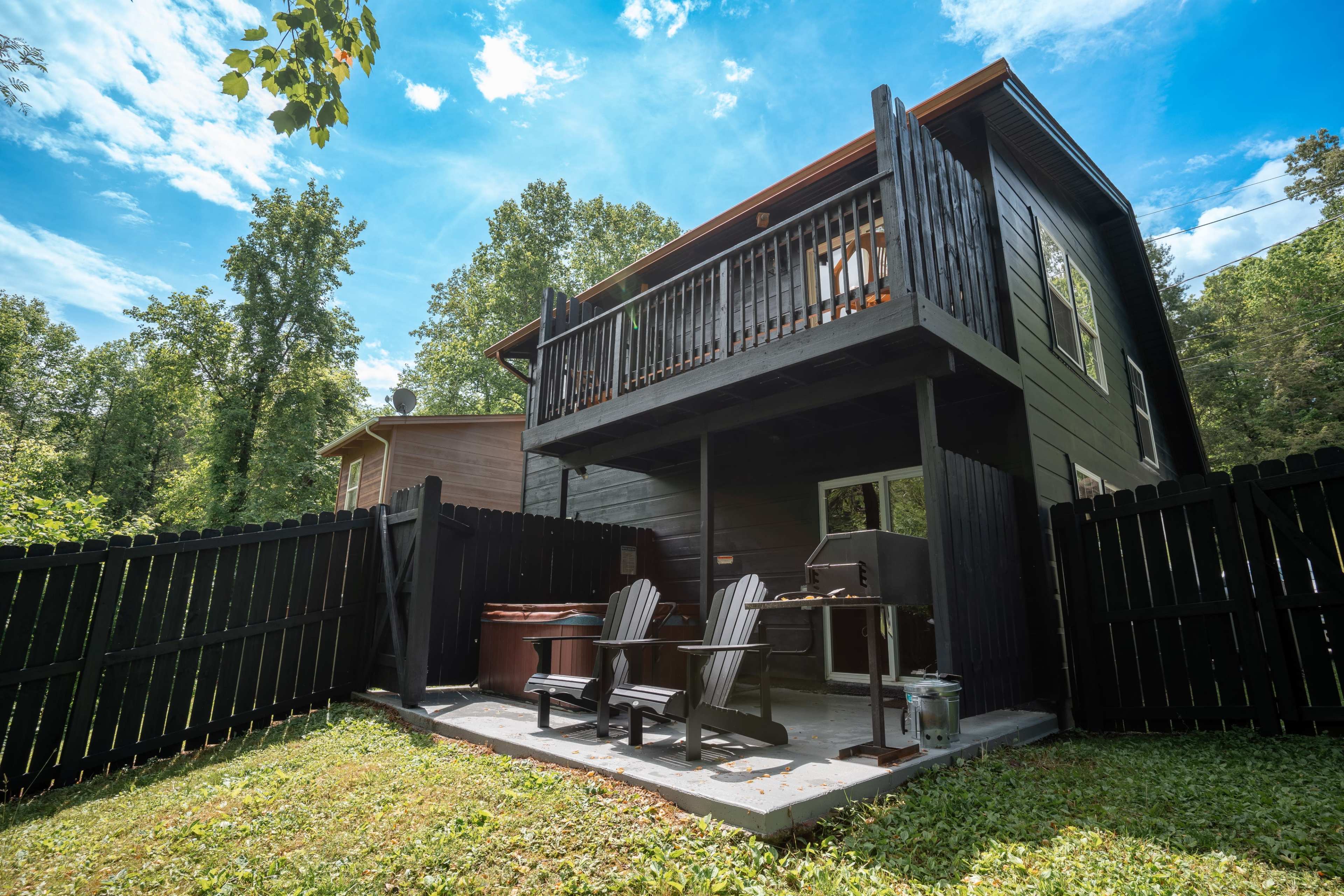 A two-story black house with a balcony overlooks a fenced outdoor area featuring seating, a grill, and a hot tub surrounded by trees.