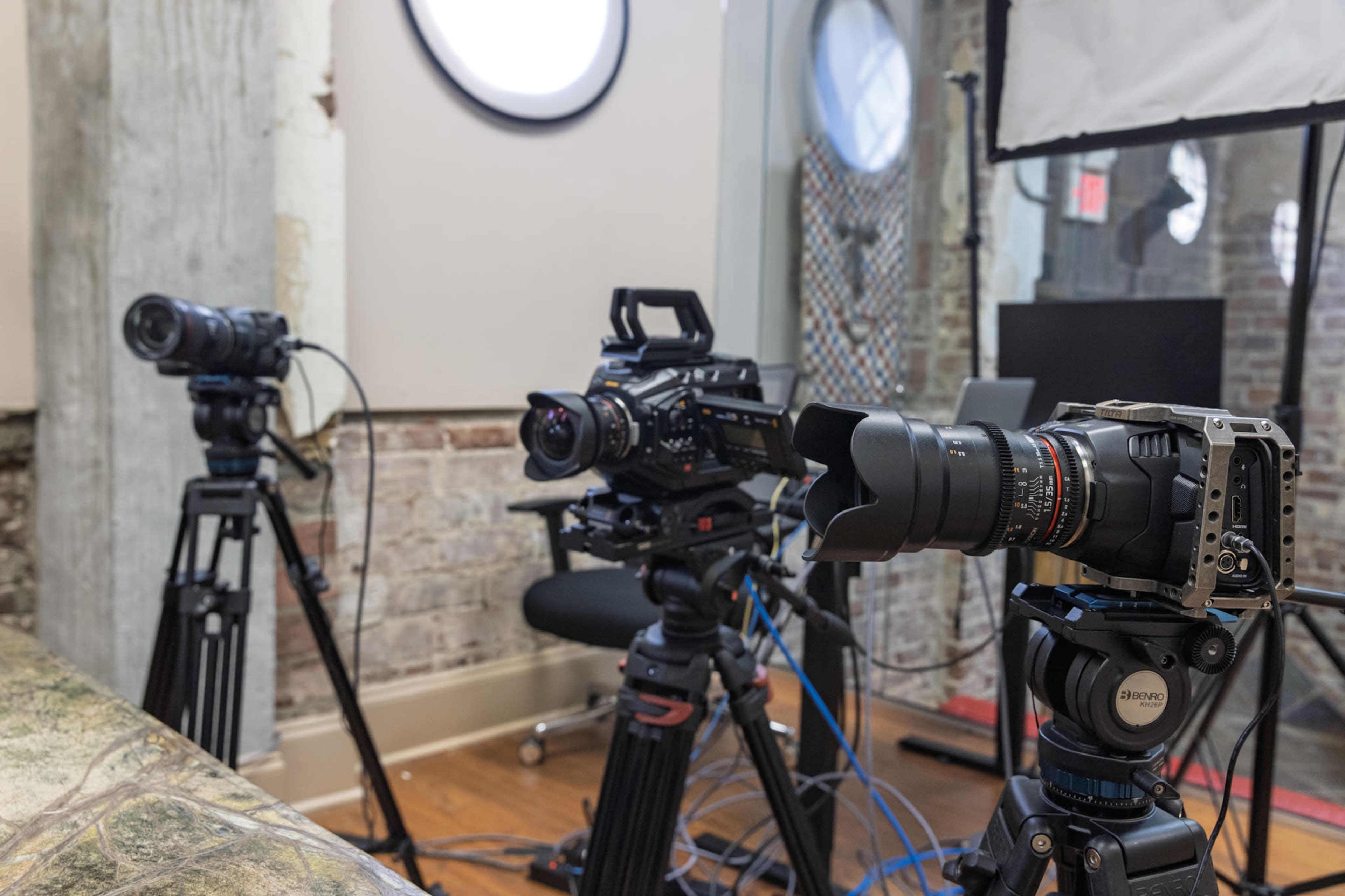Three cameras are set up on tripods in a studio environment, with a brick wall and various equipment visible in the background.