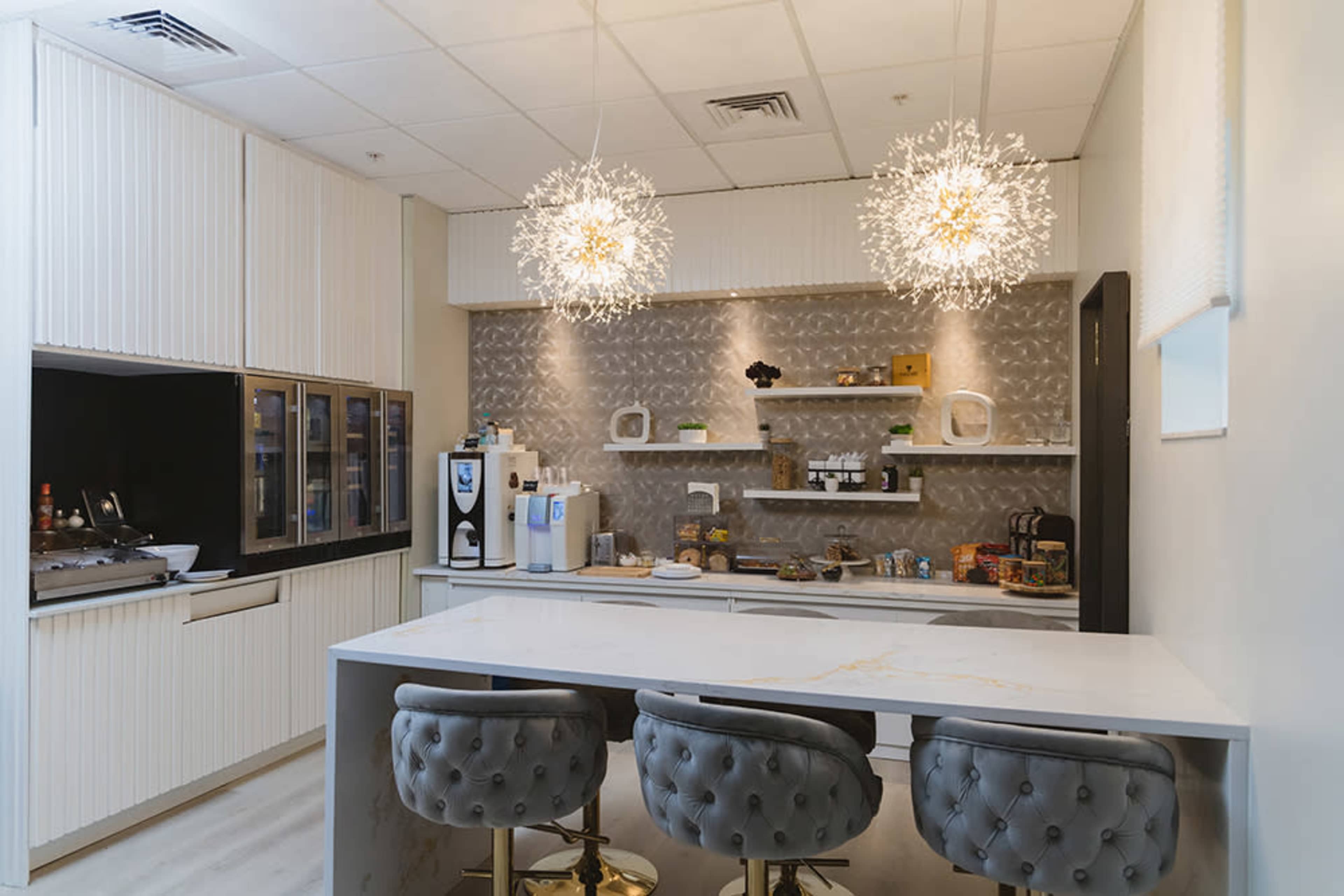 A modern kitchen space featuring a white countertop with three bar stools, a beverage station with appliances, and two decorative light fixtures hanging above.
