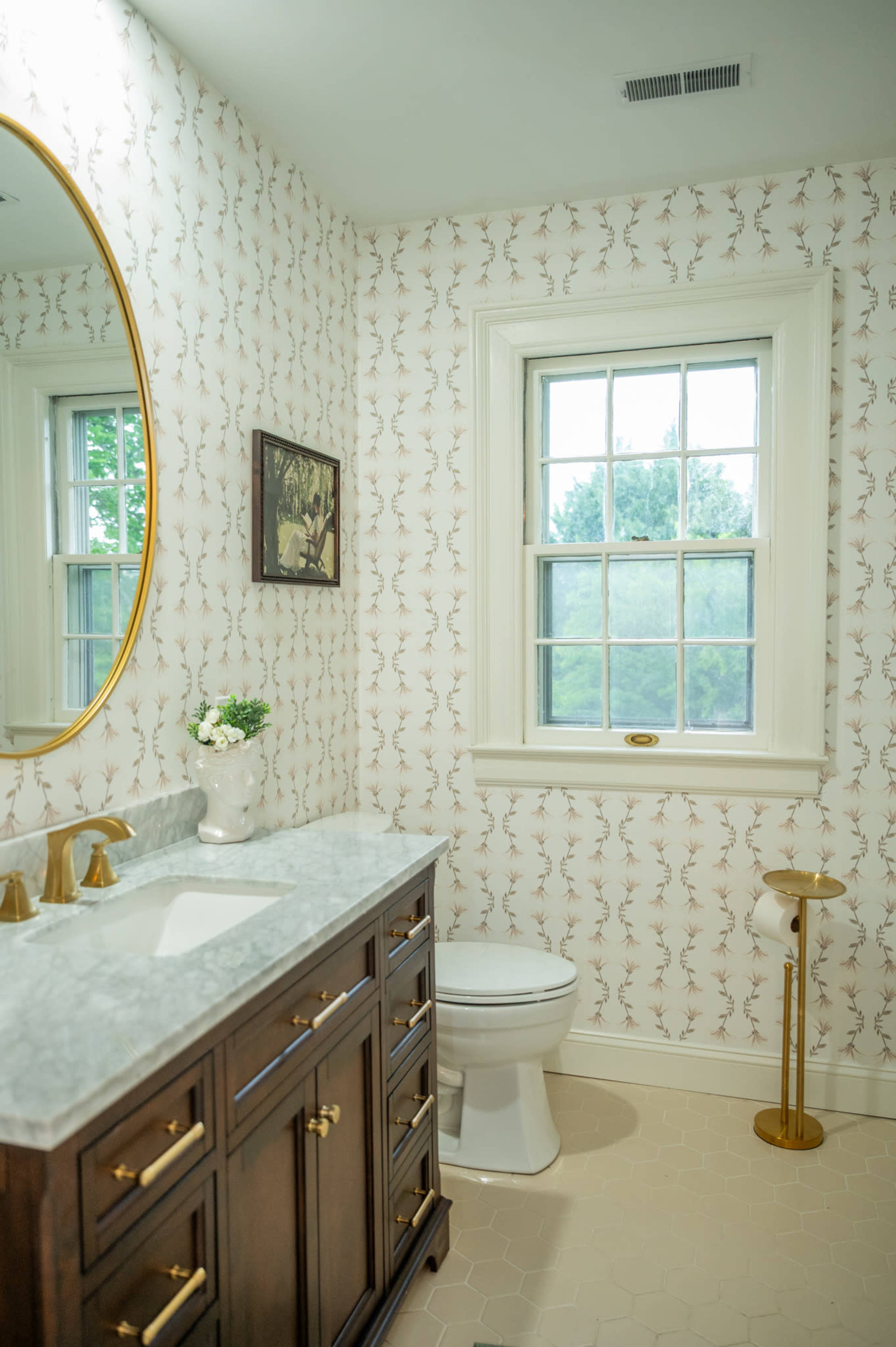 The image shows a bathroom featuring a wooden vanity with a marble countertop, a round gold mirror, and a white toilet, along with a window and floral wallpaper.