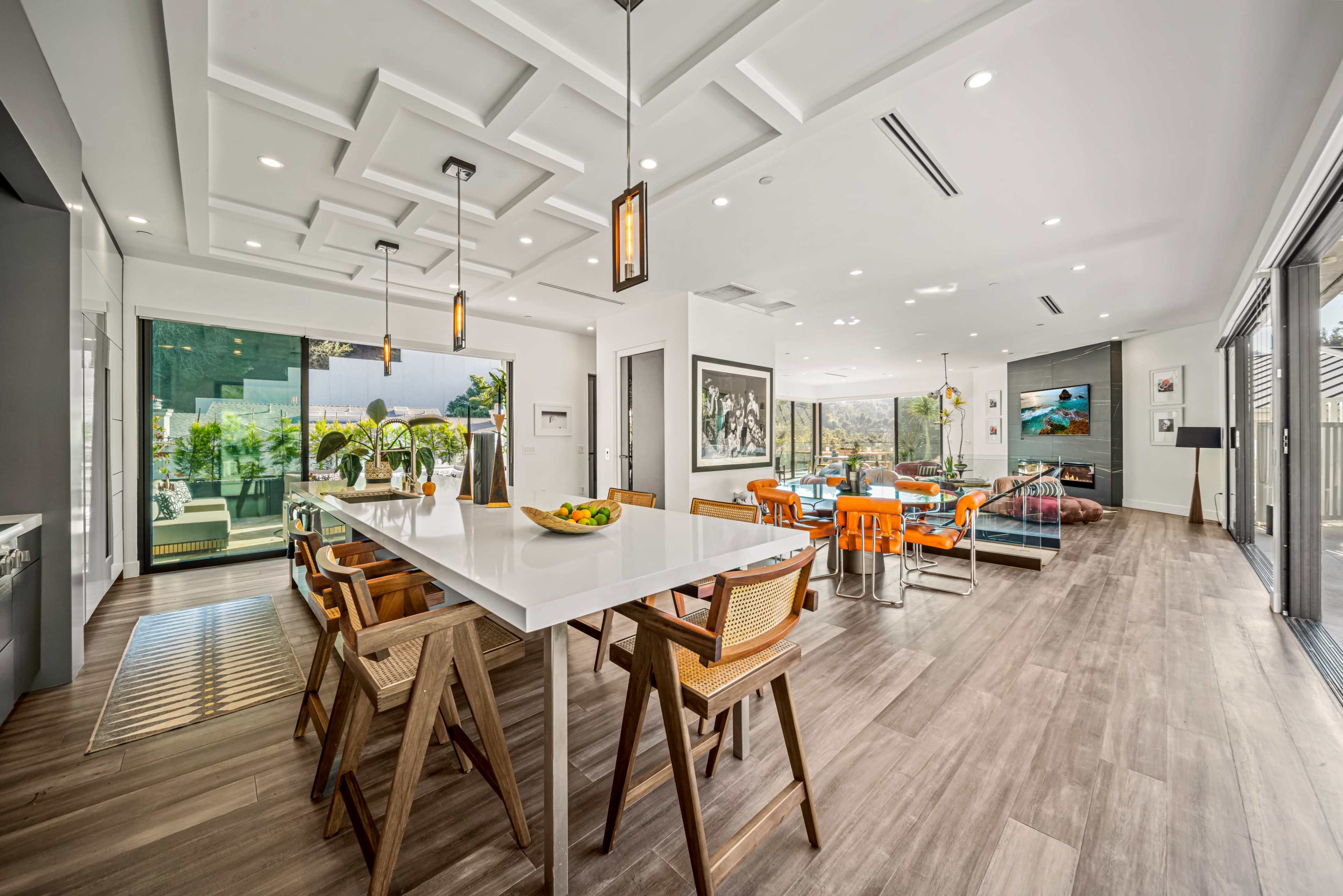 The image shows a modern dining area and open-concept living space with large windows, a long white table, wooden chairs, and vibrant decor.