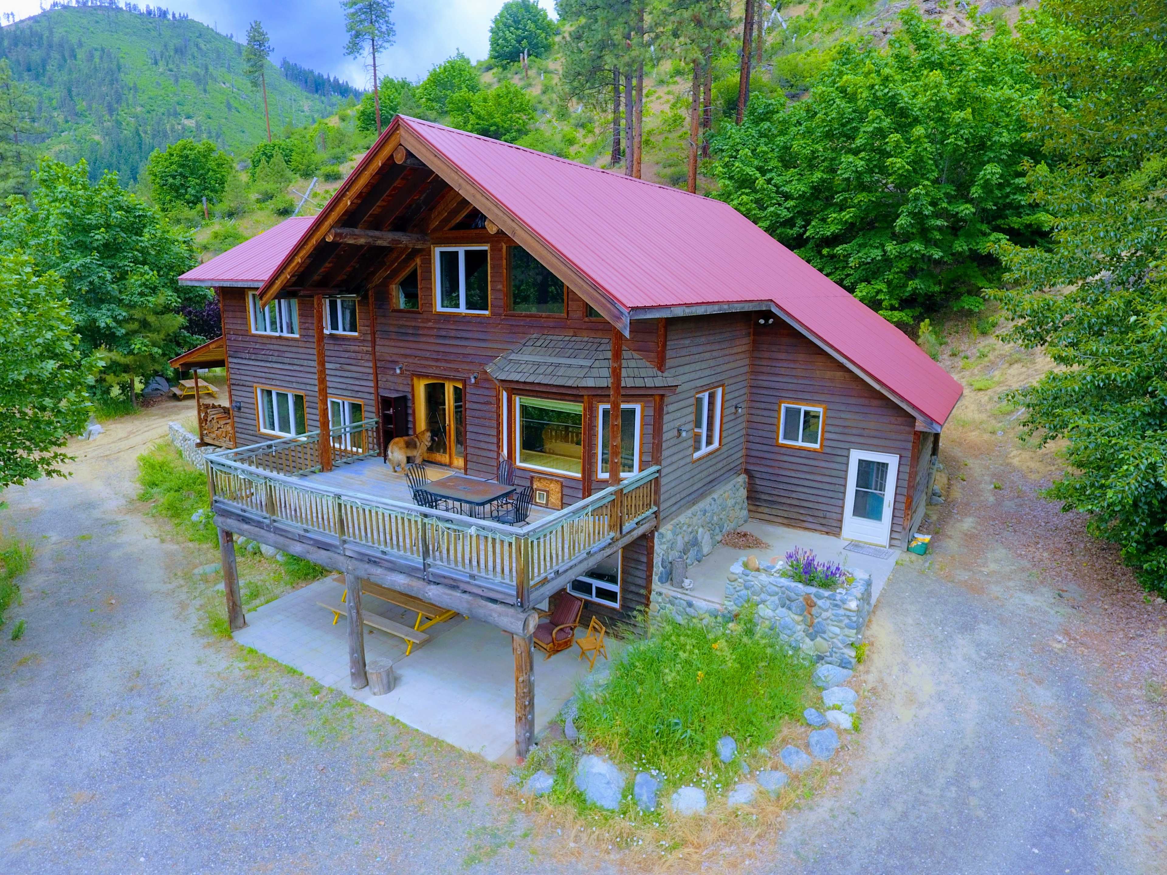 A large, two-story wooden house with a red metal roof features a spacious deck and is surrounded by trees and gravel pathways.