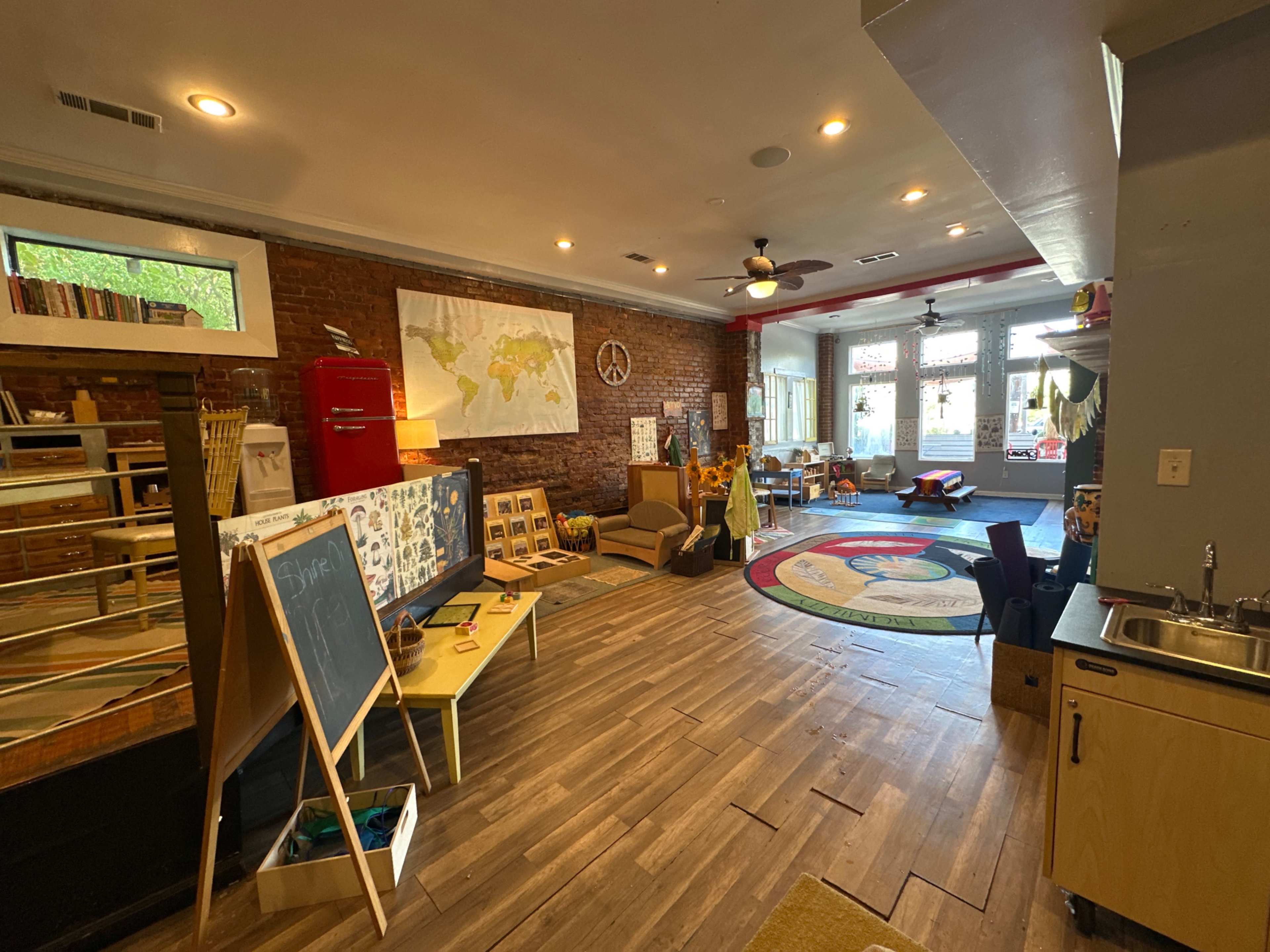 The room features wood flooring and exposed brick walls, with areas for play, learning materials, a map on the wall, and a small kitchenette.