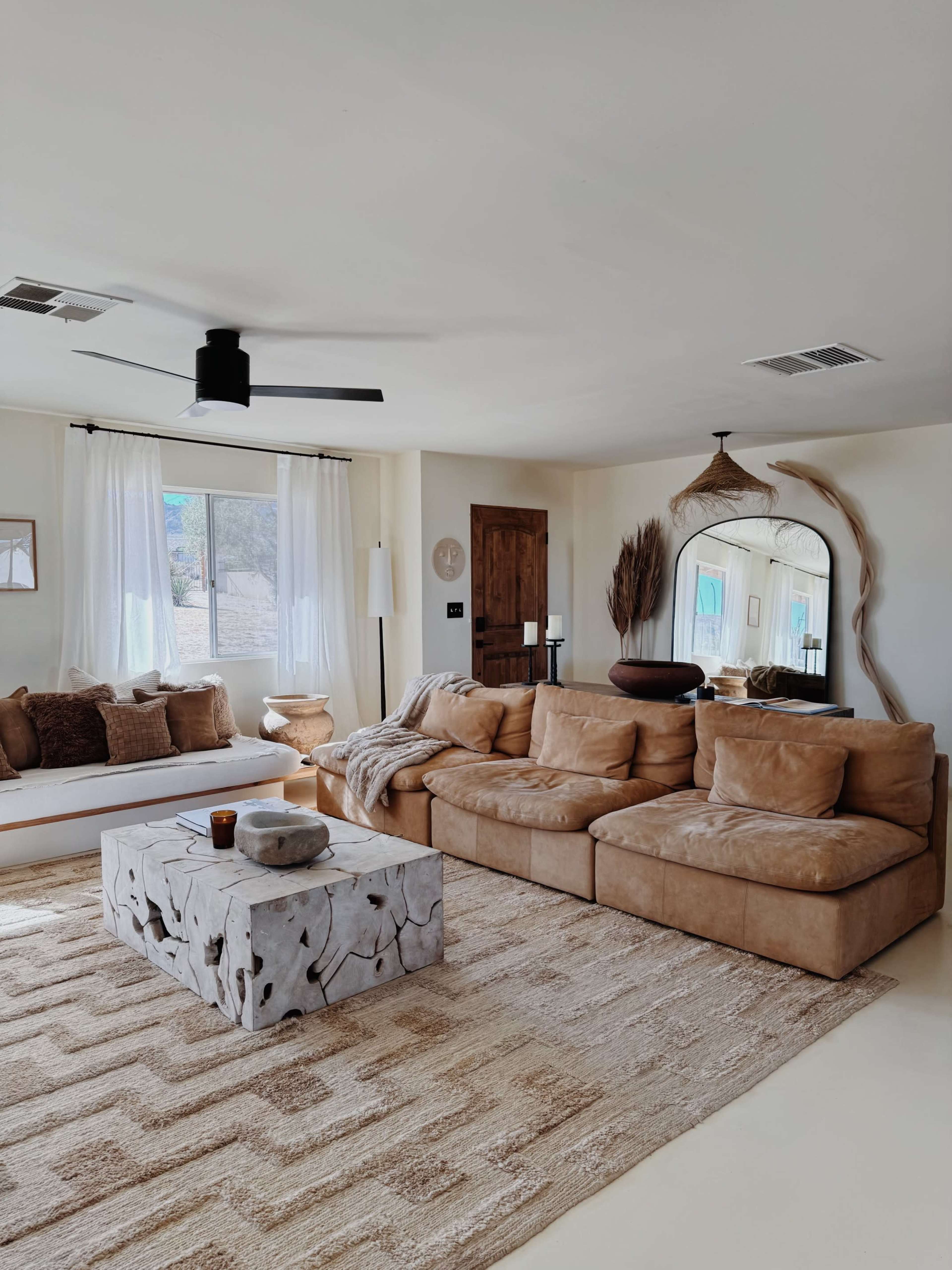 The image shows a cozy living room featuring a large sectional sofa, a textured rug, a minimalist coffee table, and natural light filtering through the windows.