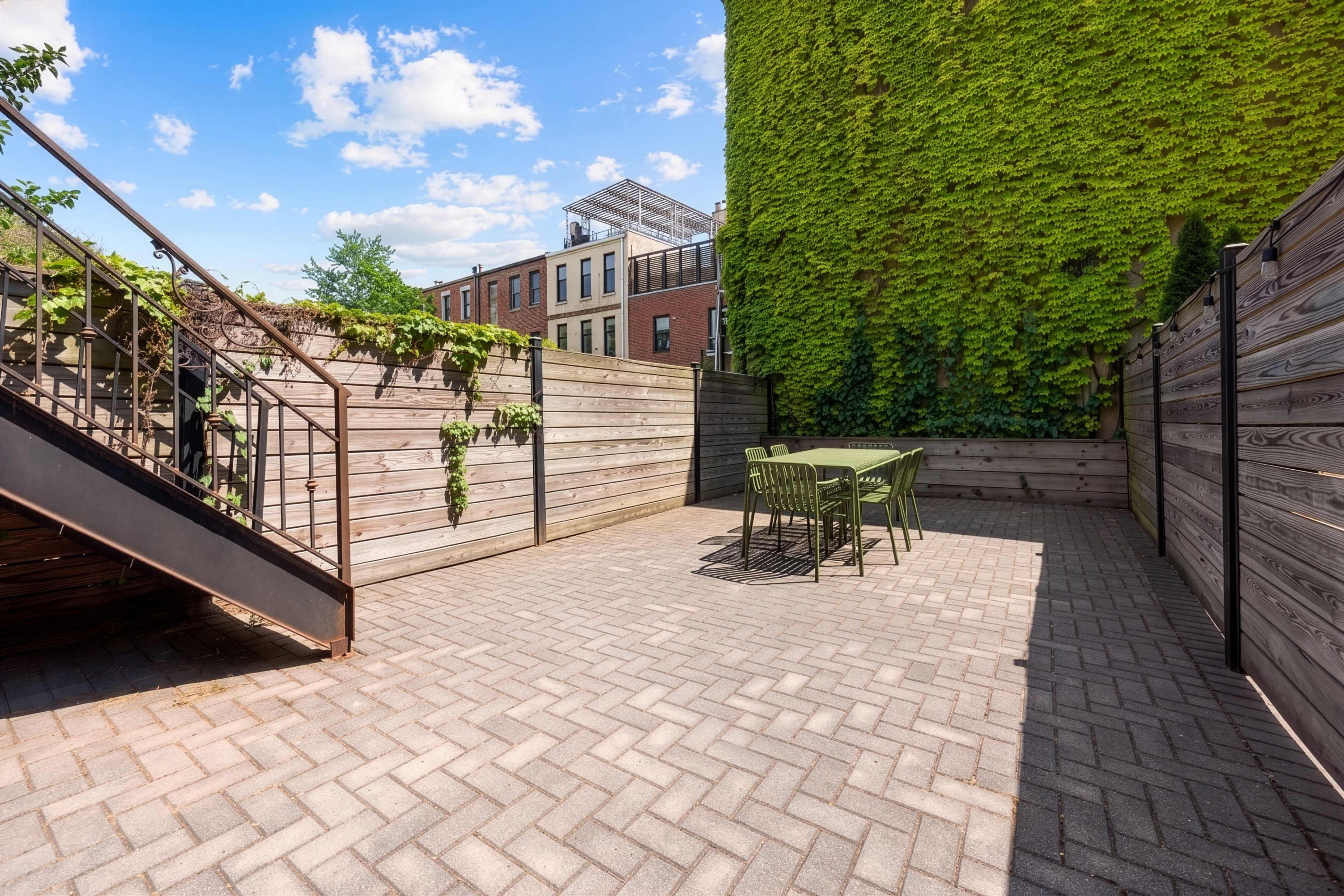 A patio area features a table and chairs surrounded by wooden fencing and a green wall of climbing plants under a blue sky with scattered clouds.