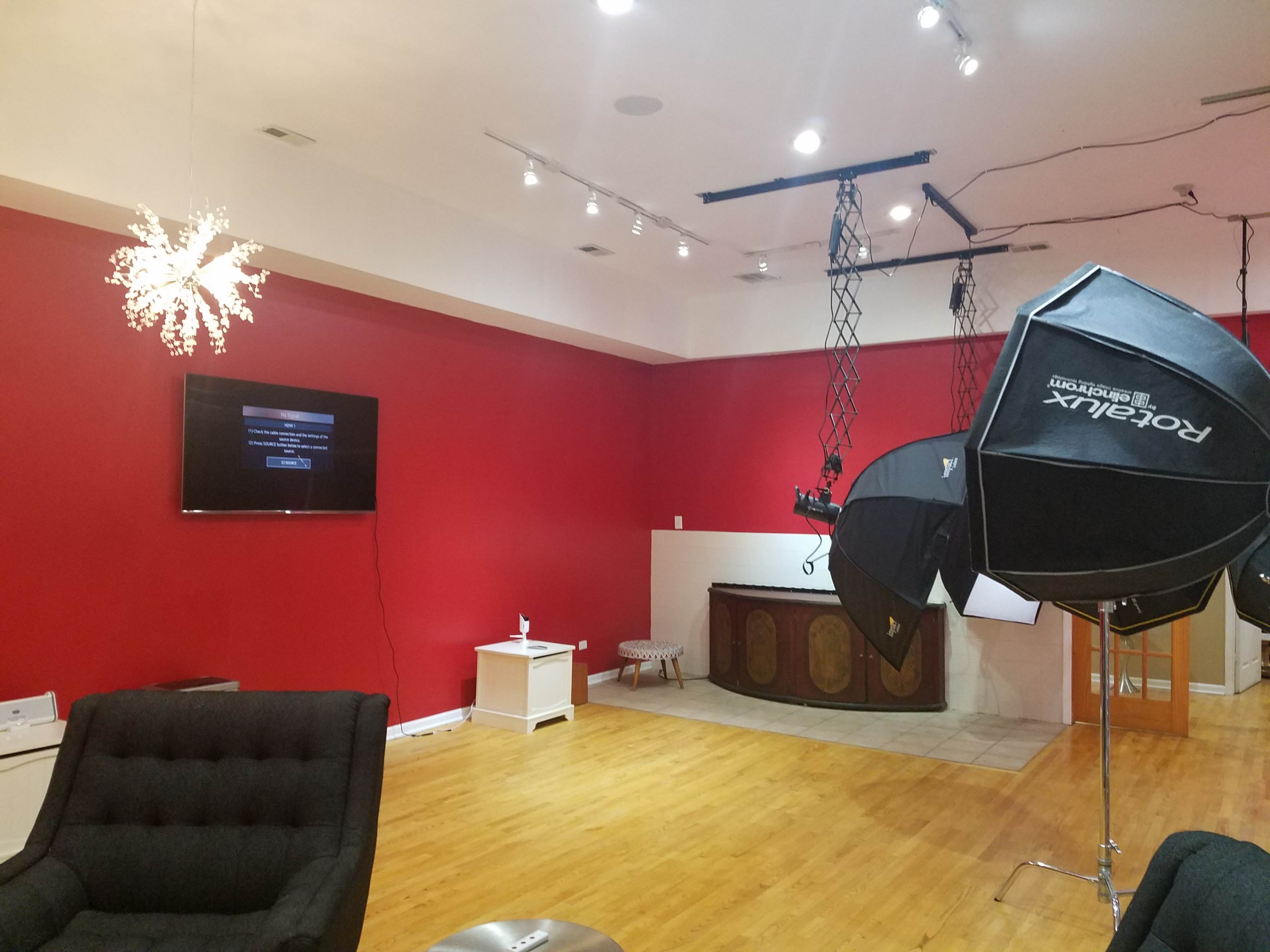 A spacious room with a red wall, a television mounted on the wall, and a lighting setup including softbox lights.
