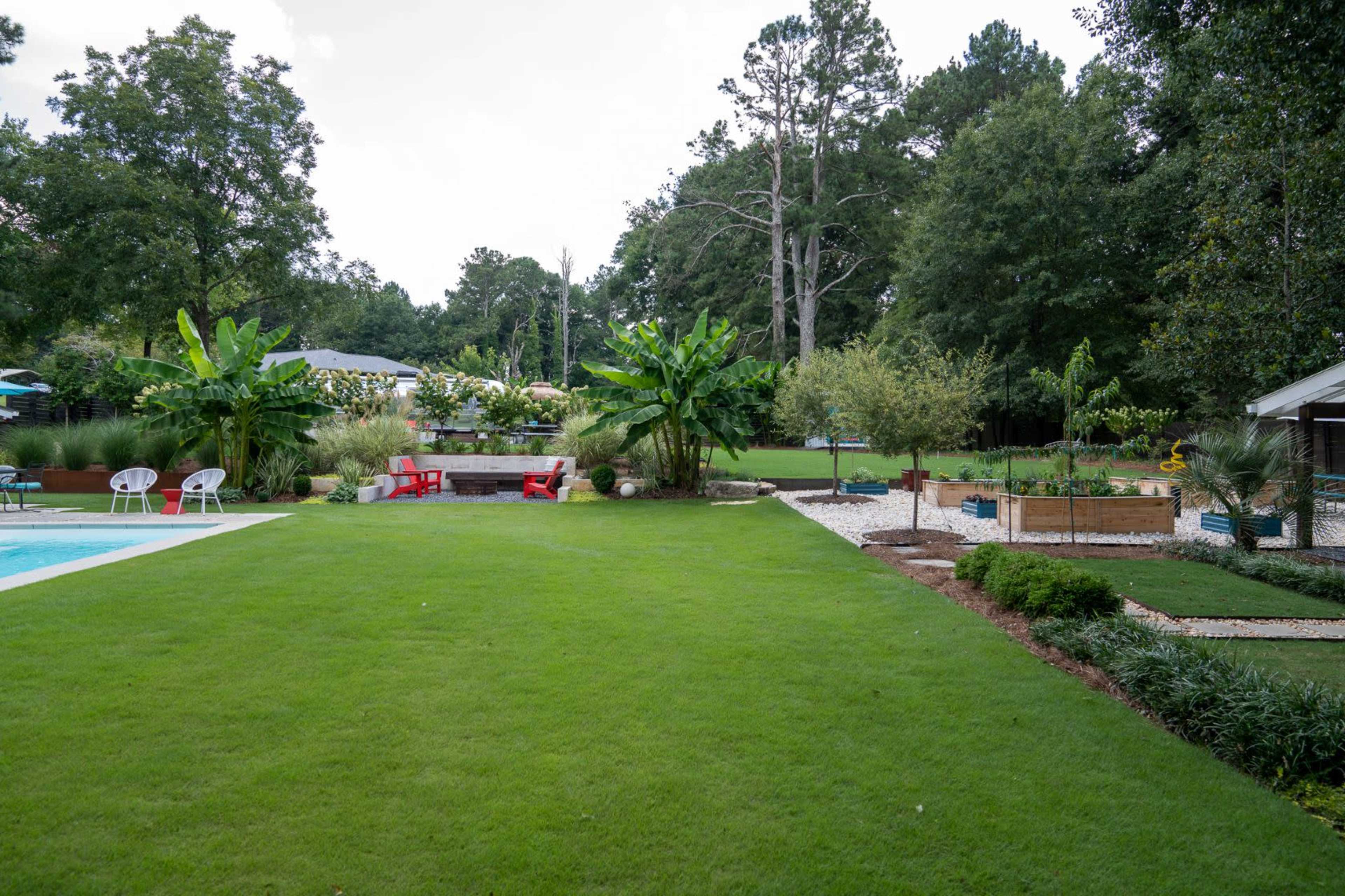The image shows a well-maintained backyard featuring a swimming pool, curated plant beds, and seating areas surrounded by lush greenery.