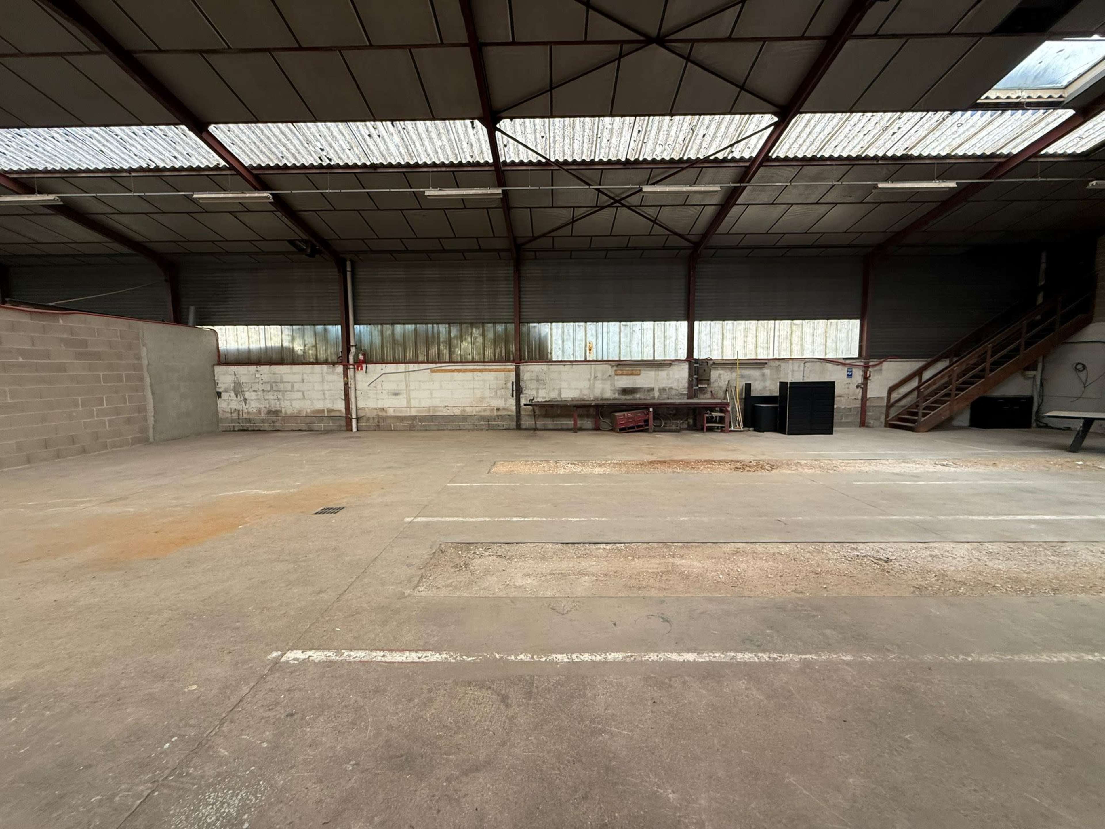 The image shows an empty warehouse with concrete flooring and metal beams, featuring a staircase and various workstations along the walls.