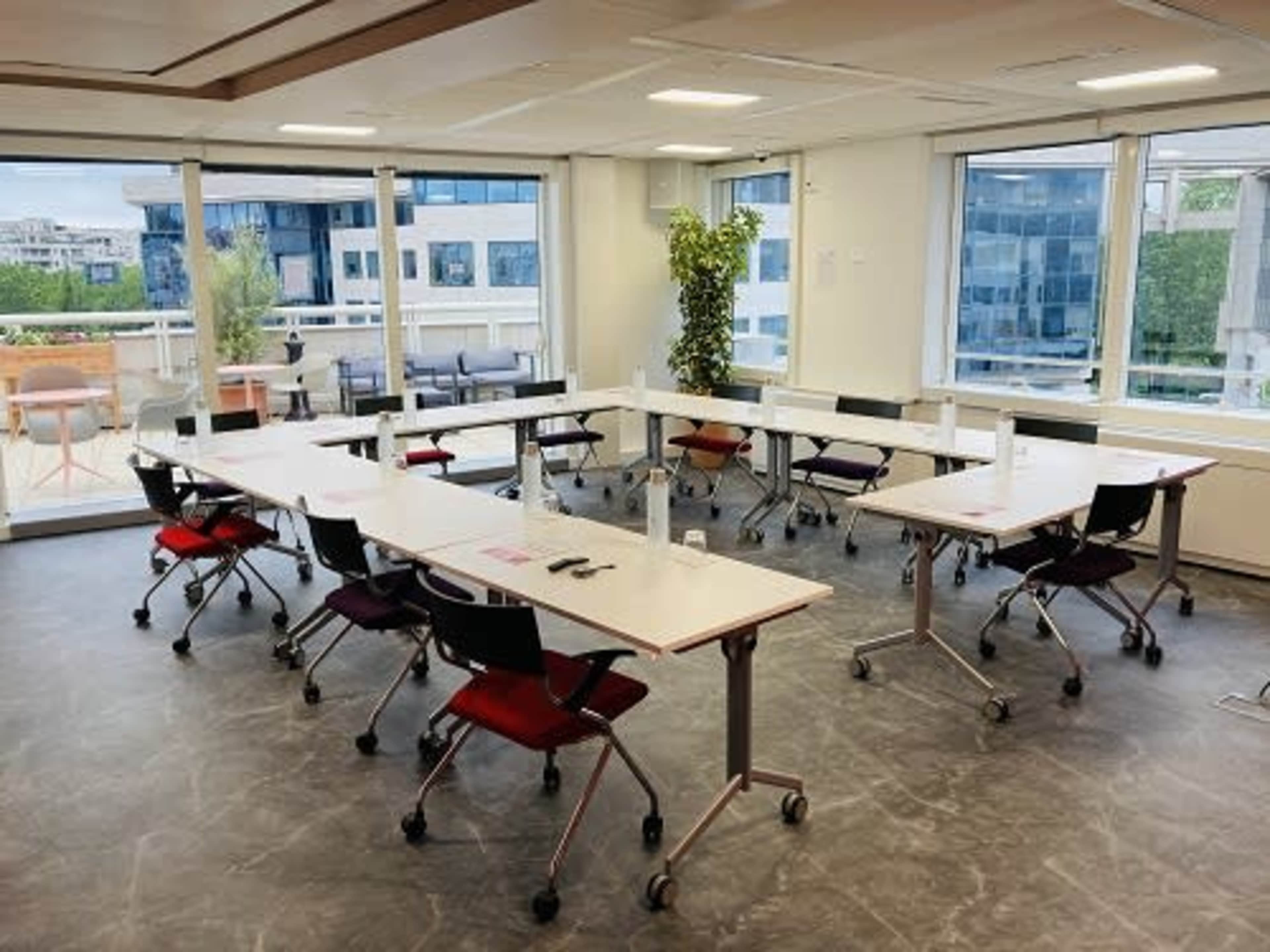 A modern meeting room features several arranged tables and chairs, with large windows overlooking an outdoor area.