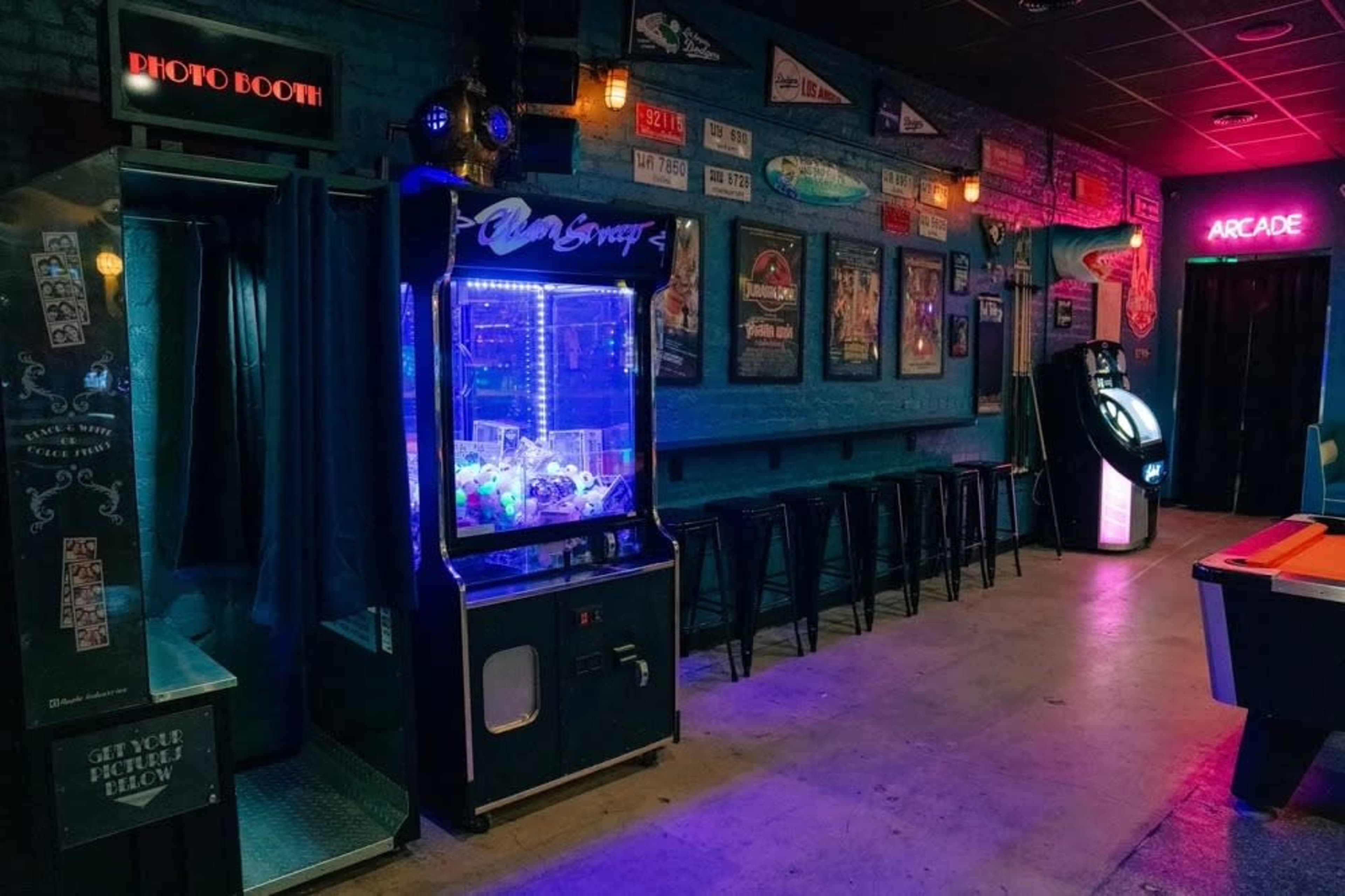 The image shows a dimly lit arcade room featuring a photo booth, a claw machine, and a row of bar stools against a blue-brick wall adorned with framed posters.