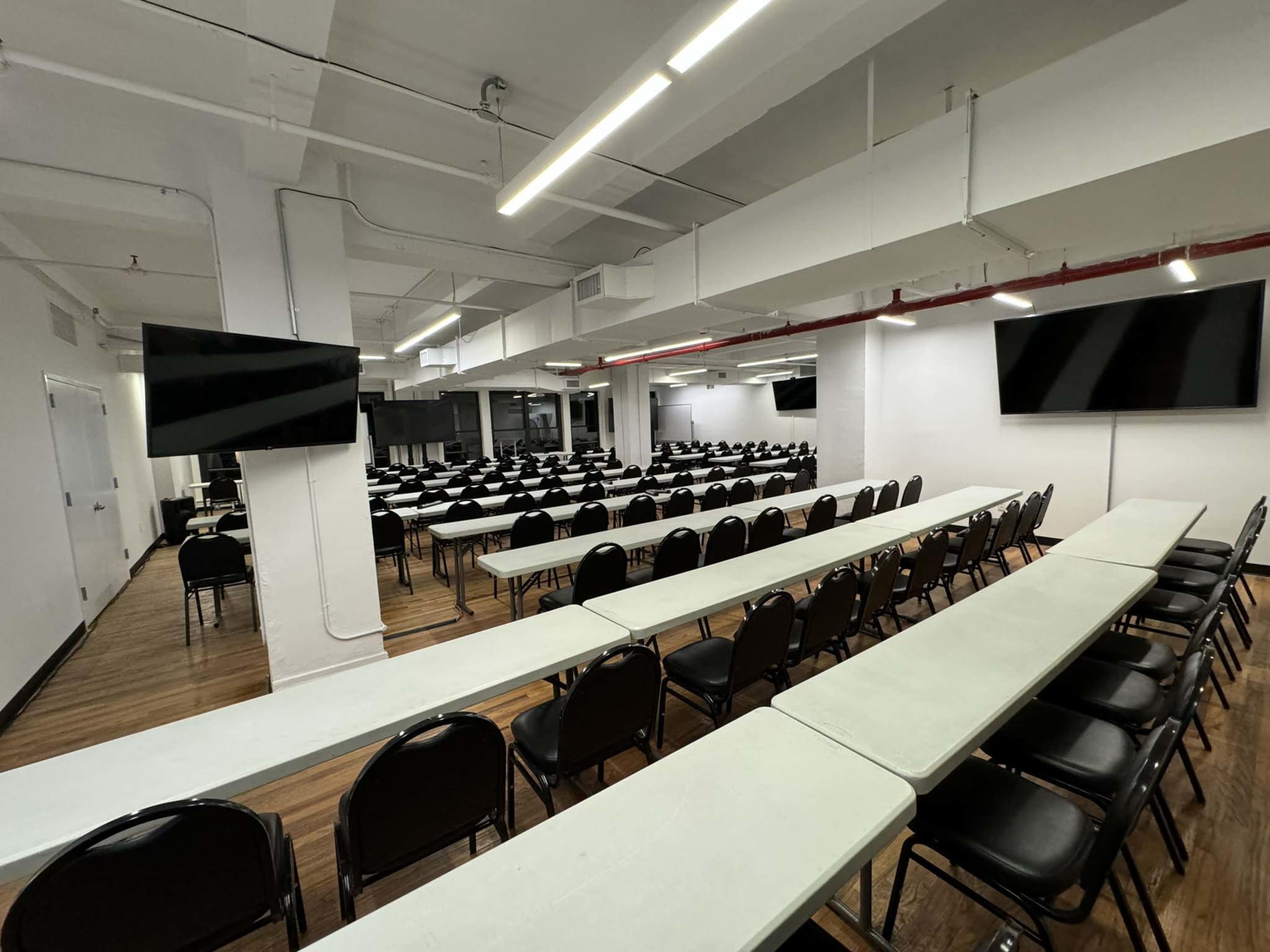 A spacious conference room is set up with rows of tables and chairs, featuring two wall-mounted screens.