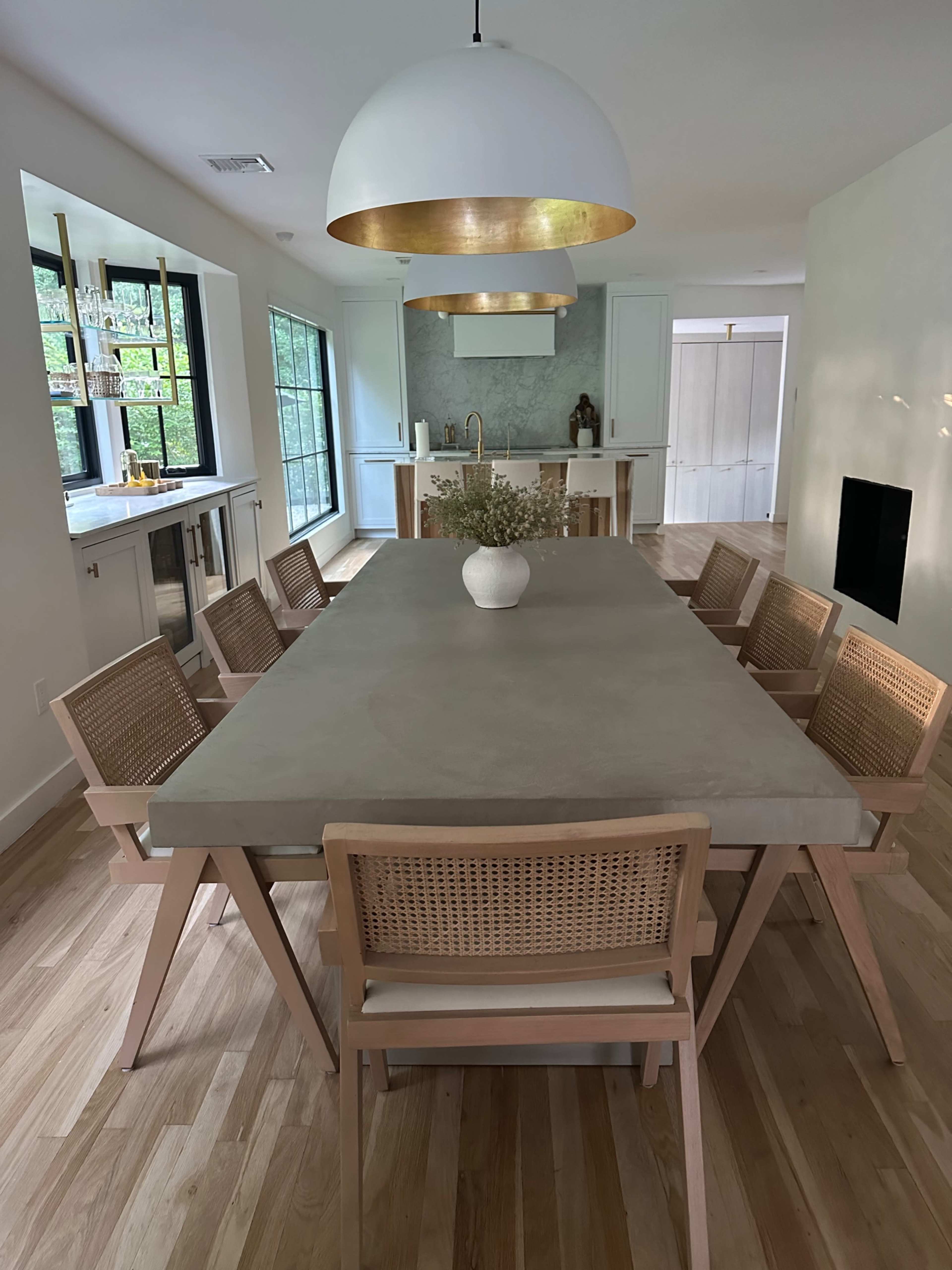 A modern dining room features a large concrete table surrounded by wooden chairs, with large windows allowing natural light to fill the space.