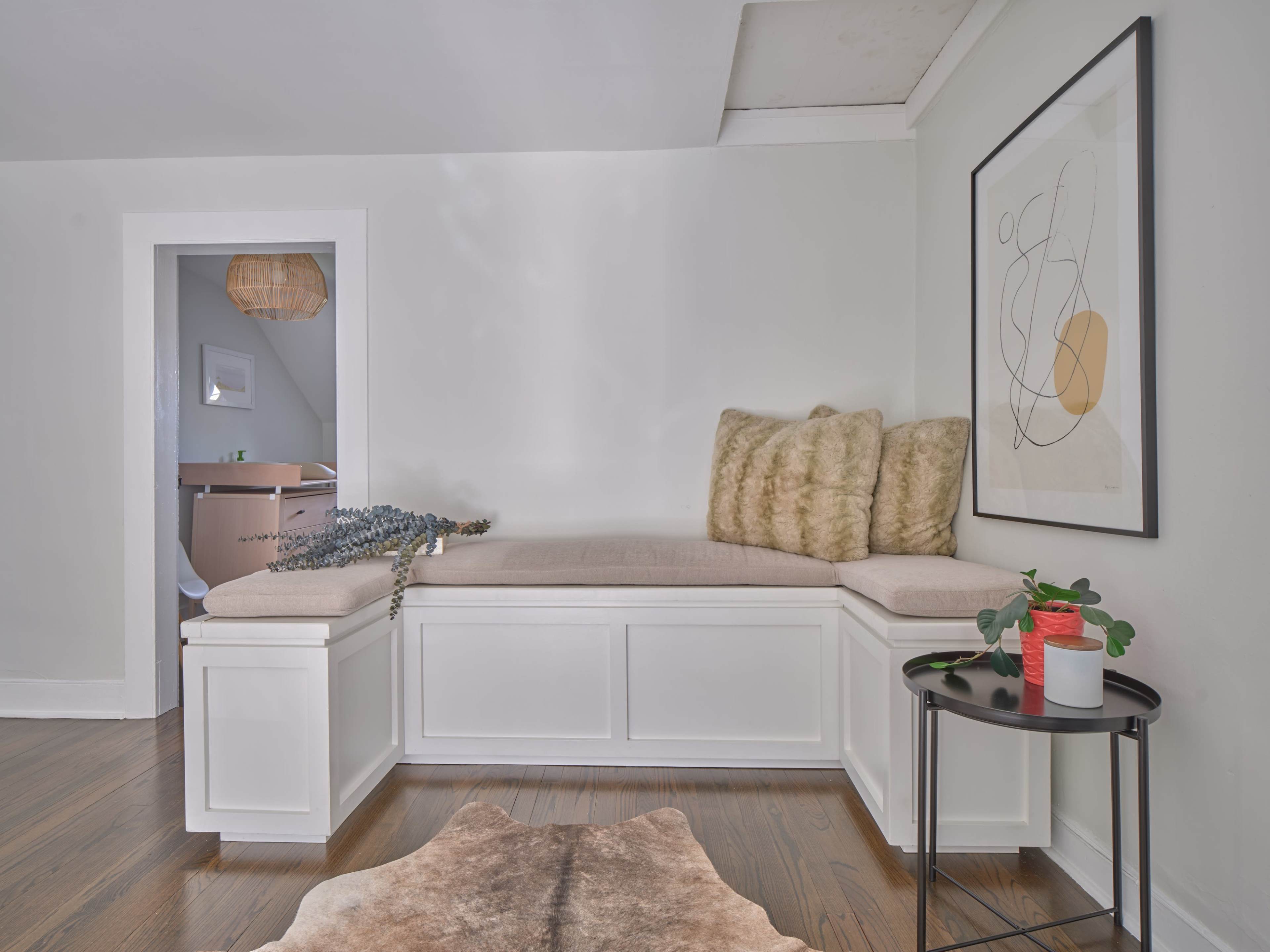 A cozy corner with a white window seat adorned with beige cushions, a small round table with a potted plant, and light wood flooring.