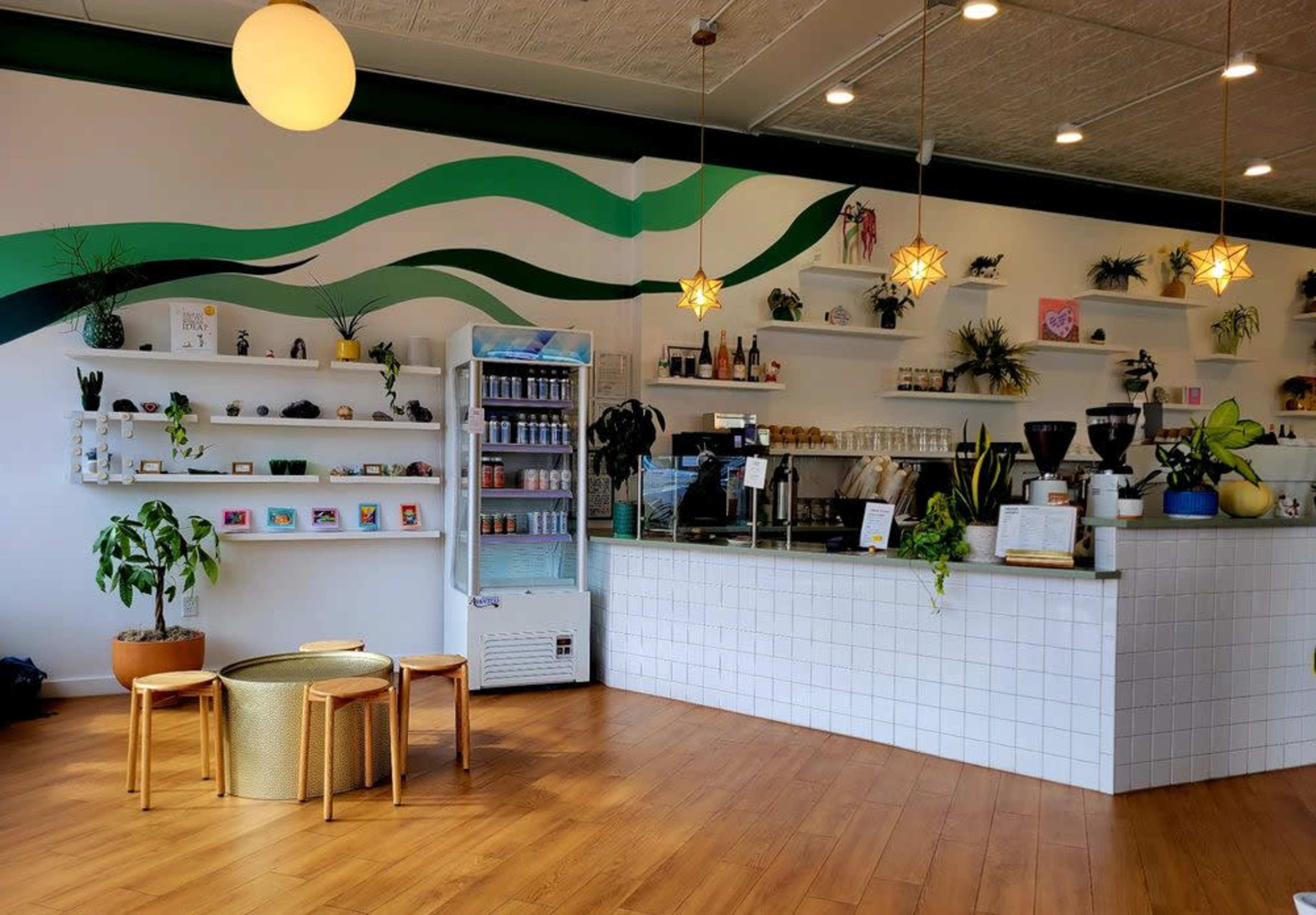 The image shows a modern coffee shop interior with a counter, shelves displaying products, seating, and a large mural featuring green waves on the wall.