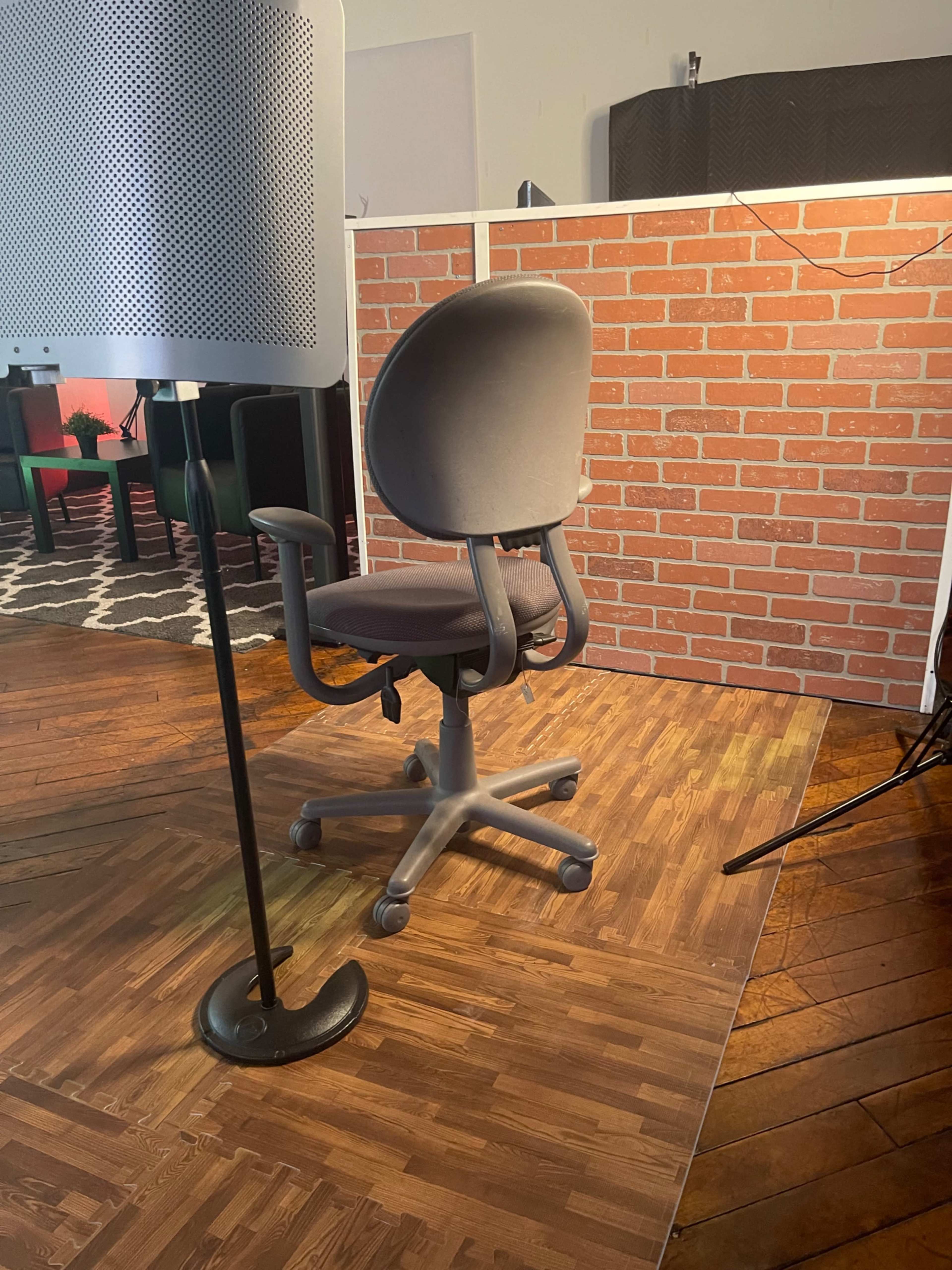 A swivel office chair is positioned on a wooden floor in front of a brick-patterned backdrop.