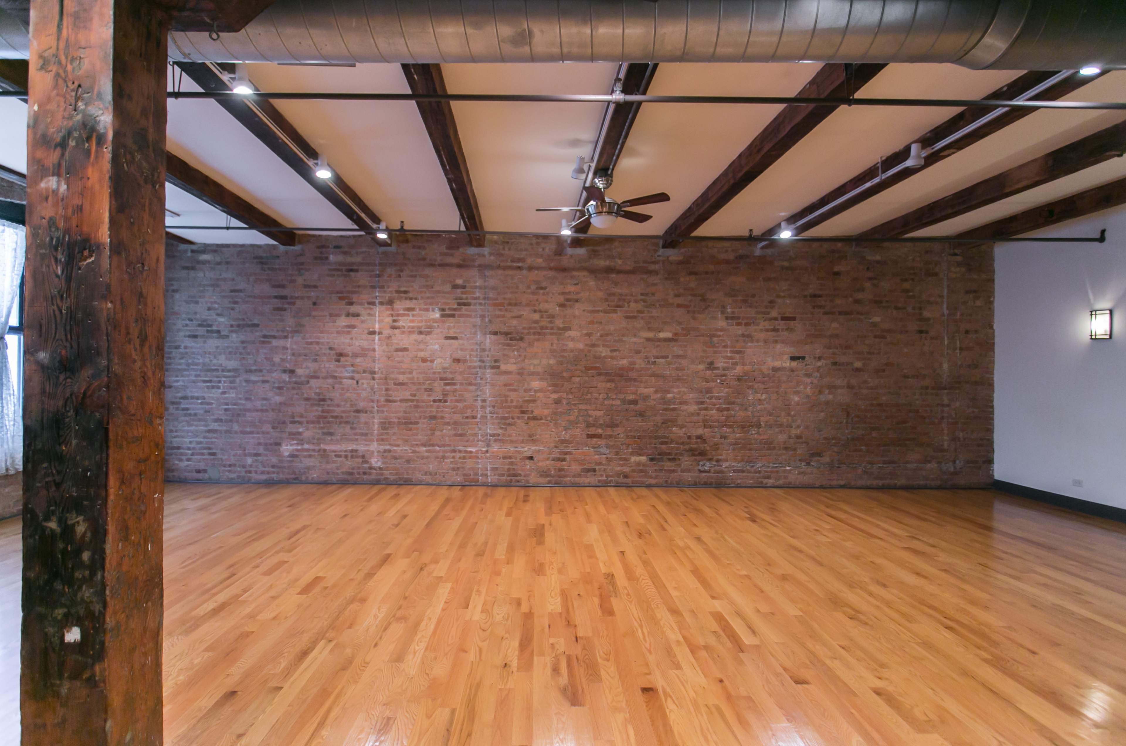 A spacious empty room with wooden flooring, exposed brick walls, and visible ceiling beams.