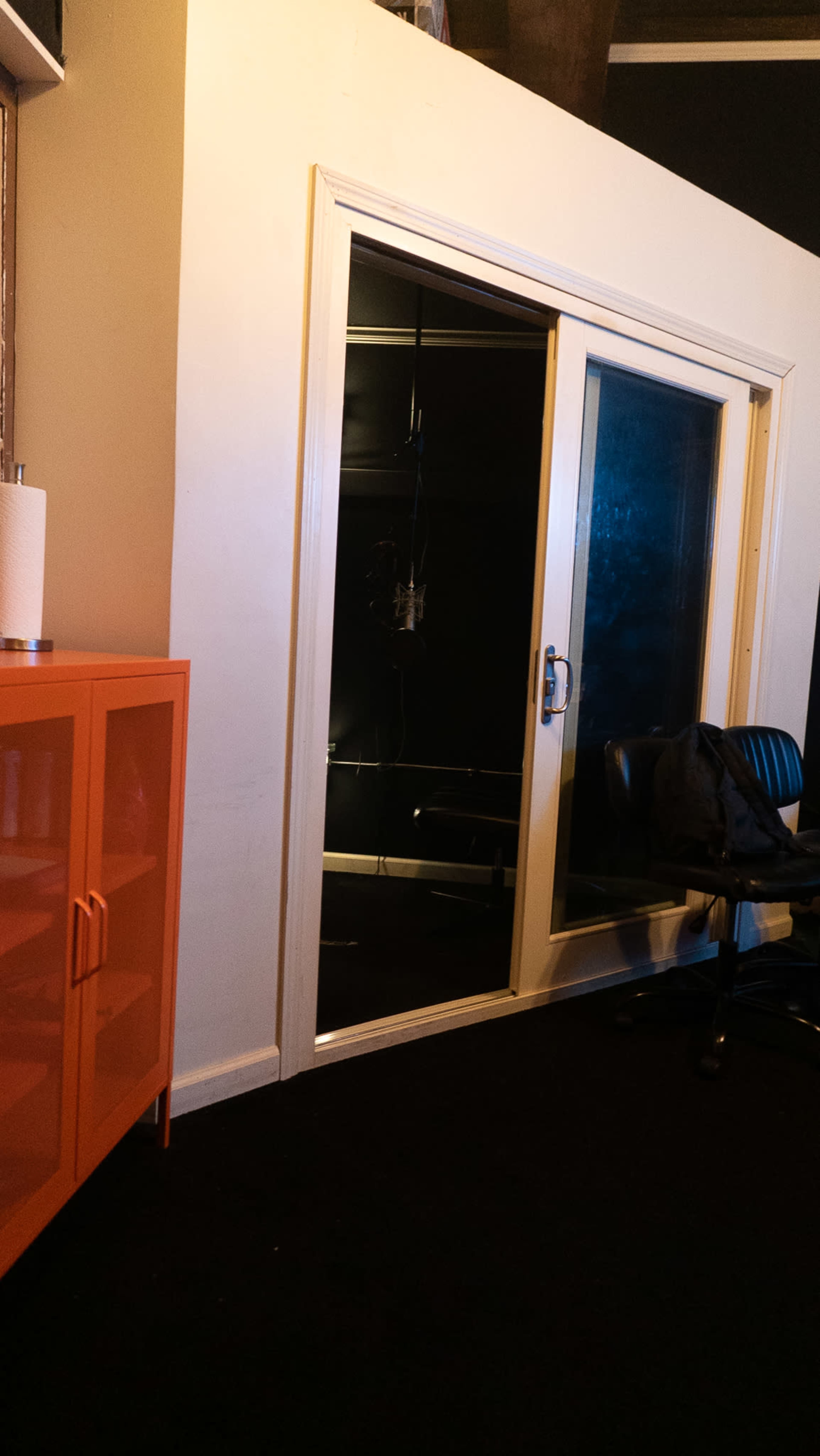 The image shows a small glass-enclosed room with a black interior, next to an orange cabinet and a black chair in a dimly lit space.
