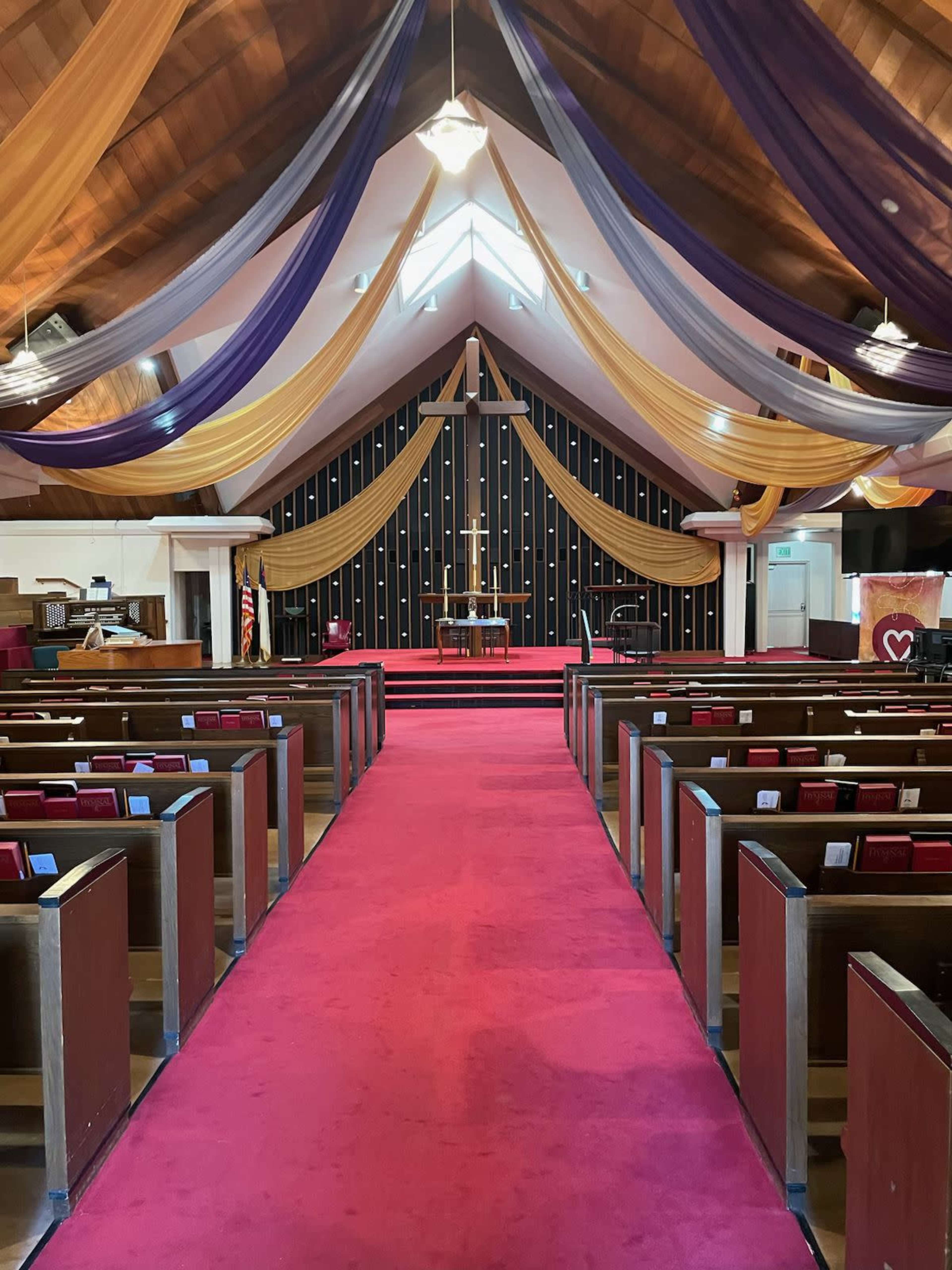 The interior of a church displays rows of wooden pews facing a stage adorned with a cross and draped fabric in purple and gold hues.