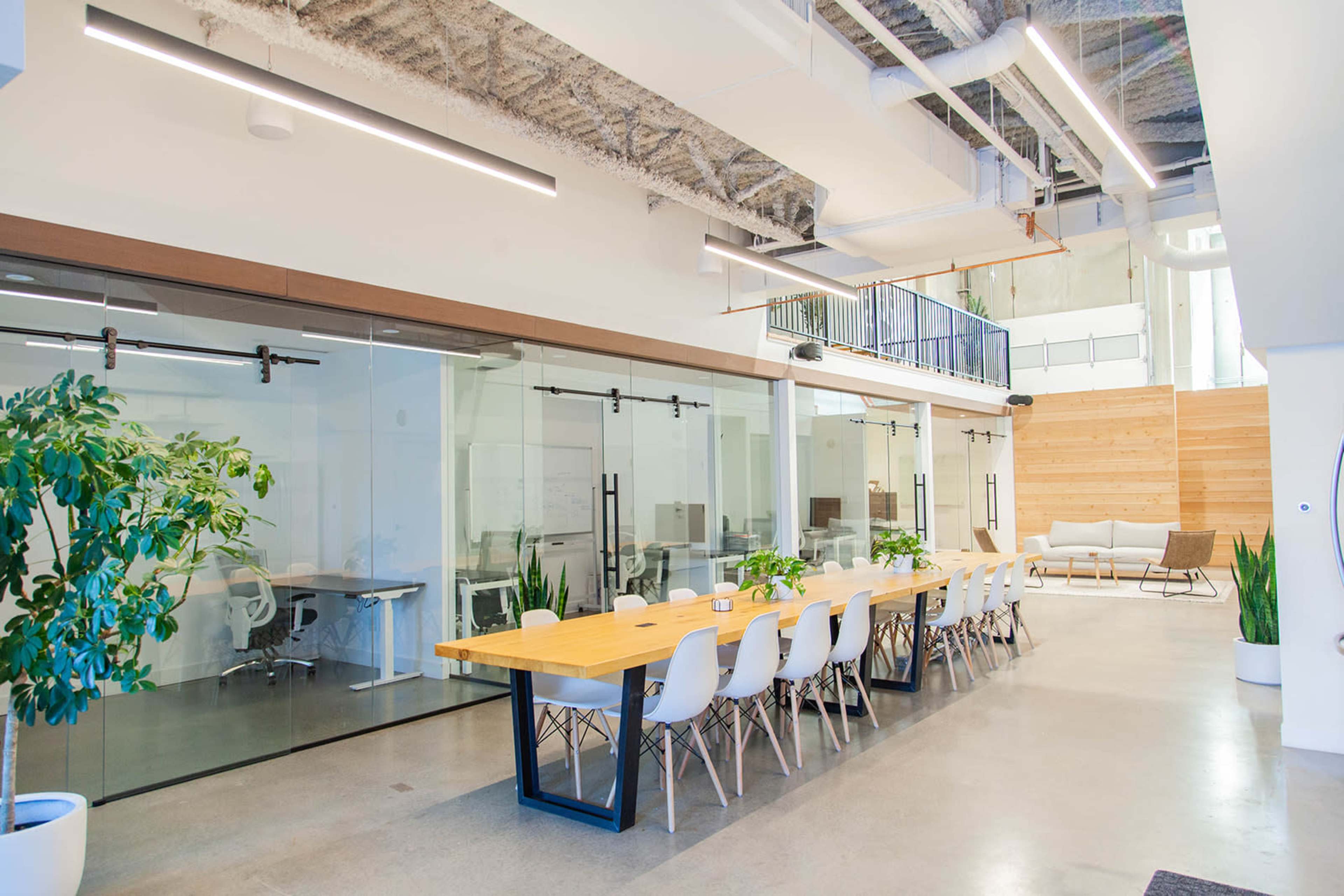 The image shows a modern office space featuring a long wooden table surrounded by white chairs, with glass-walled meeting rooms and plants lining the area.