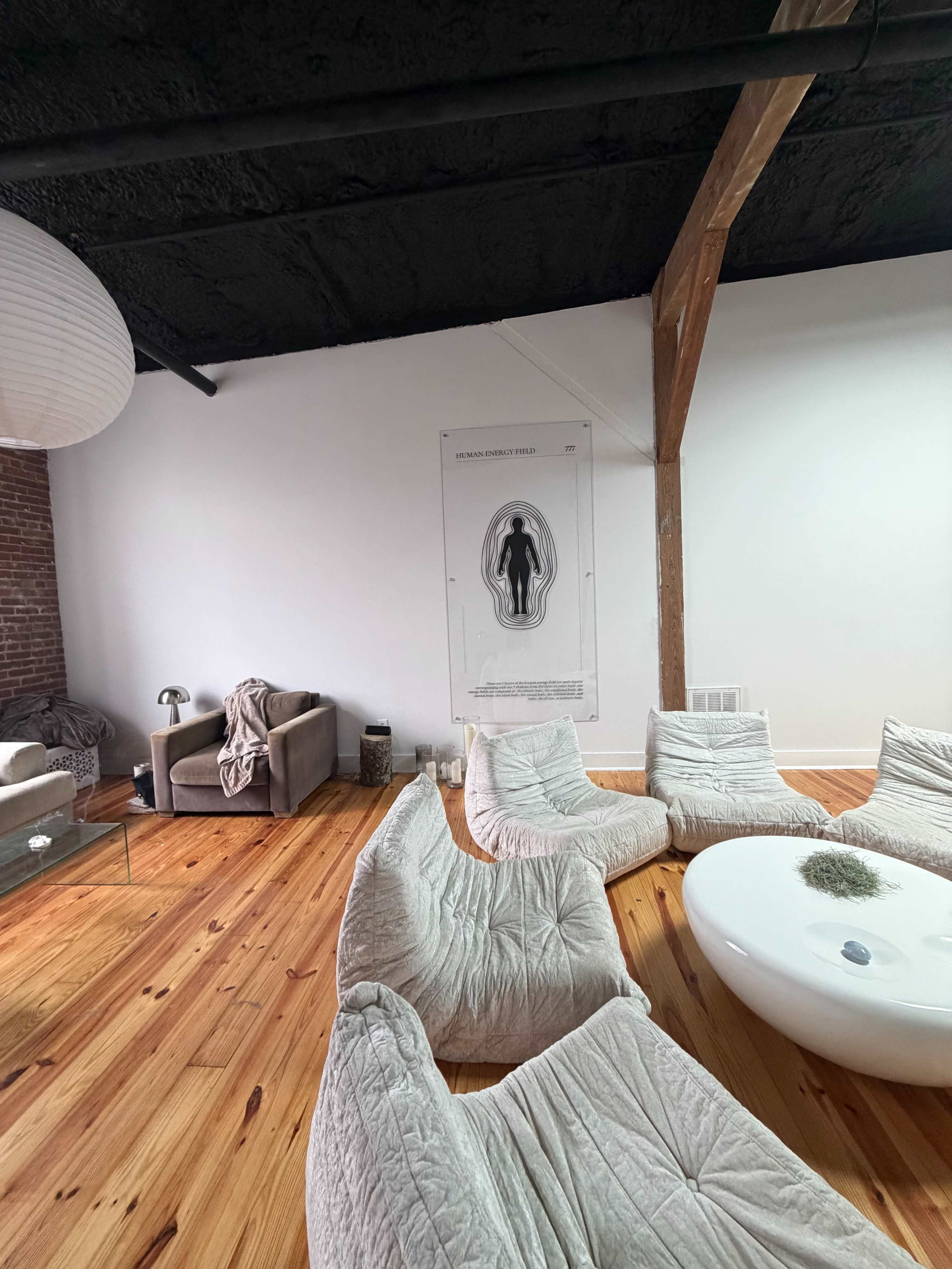 The image shows a spacious, minimalist room with wooden flooring, two armchairs, and a large poster on the wall depicting a figure within an outline.