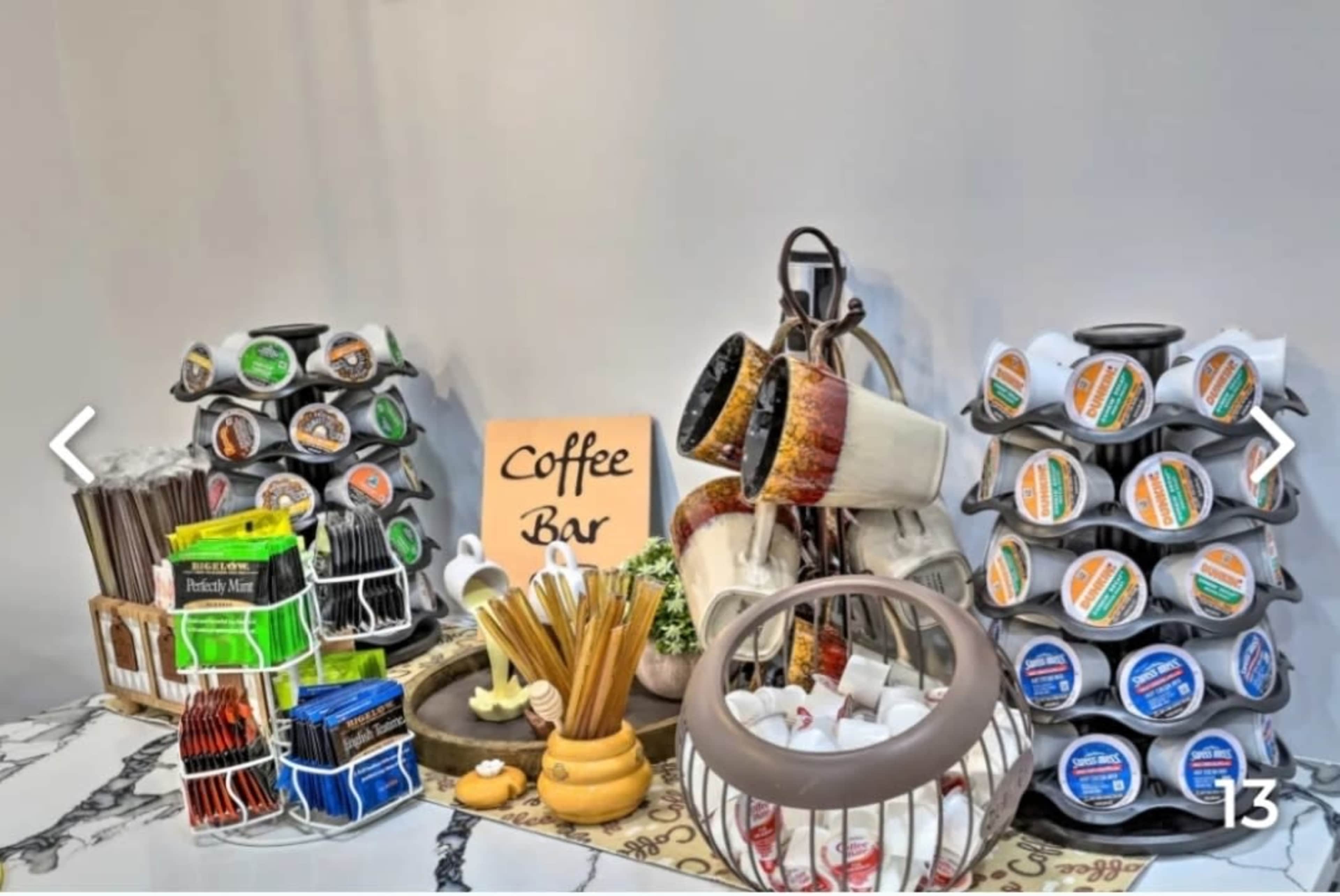 The image shows a neatly arranged coffee bar with various coffee pods, mugs, stirrers, and utensils displayed on a countertop.