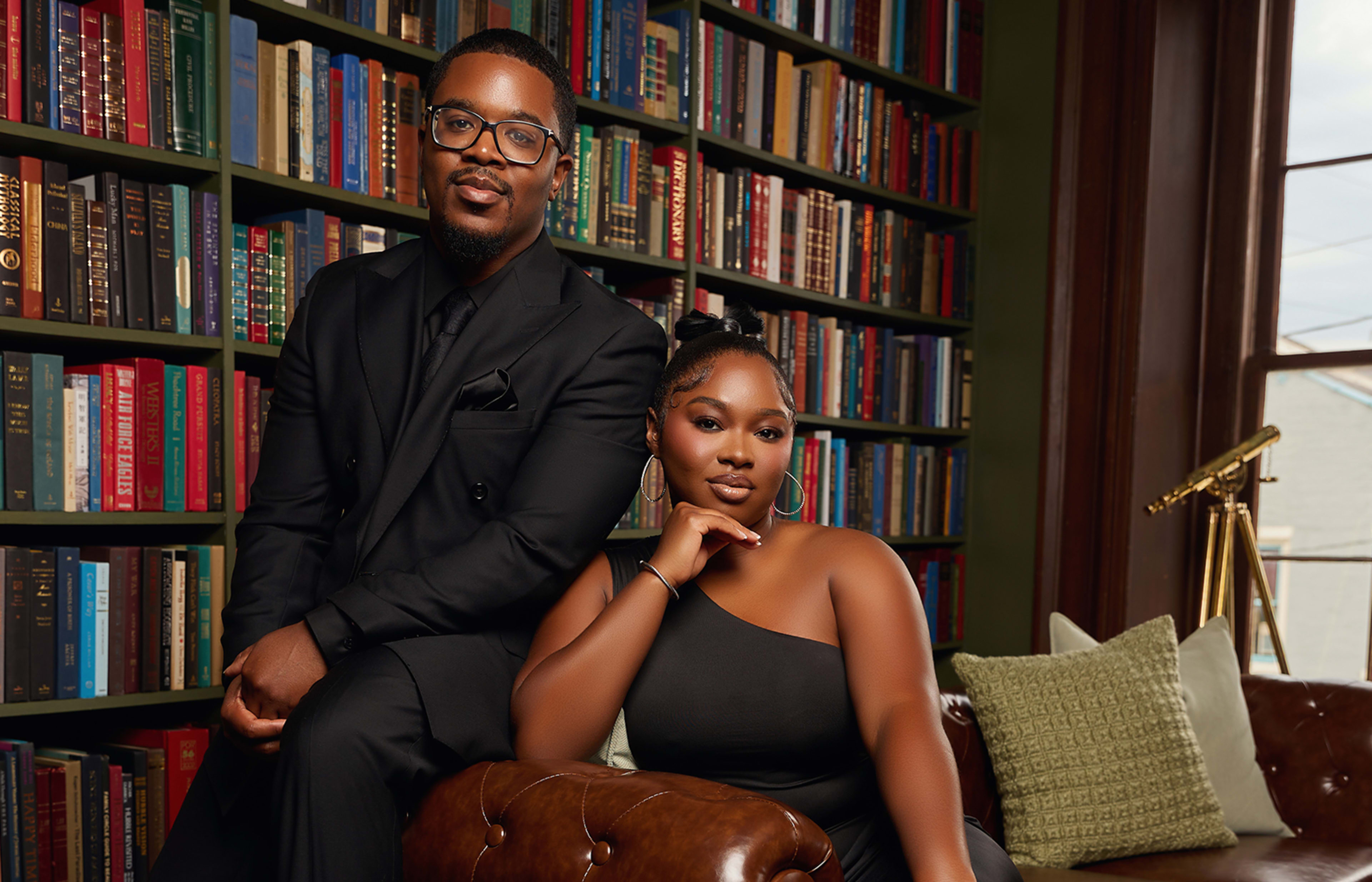 A man in a black suit and a woman in a black dress pose together in a cozy room filled with colorful books on shelves.