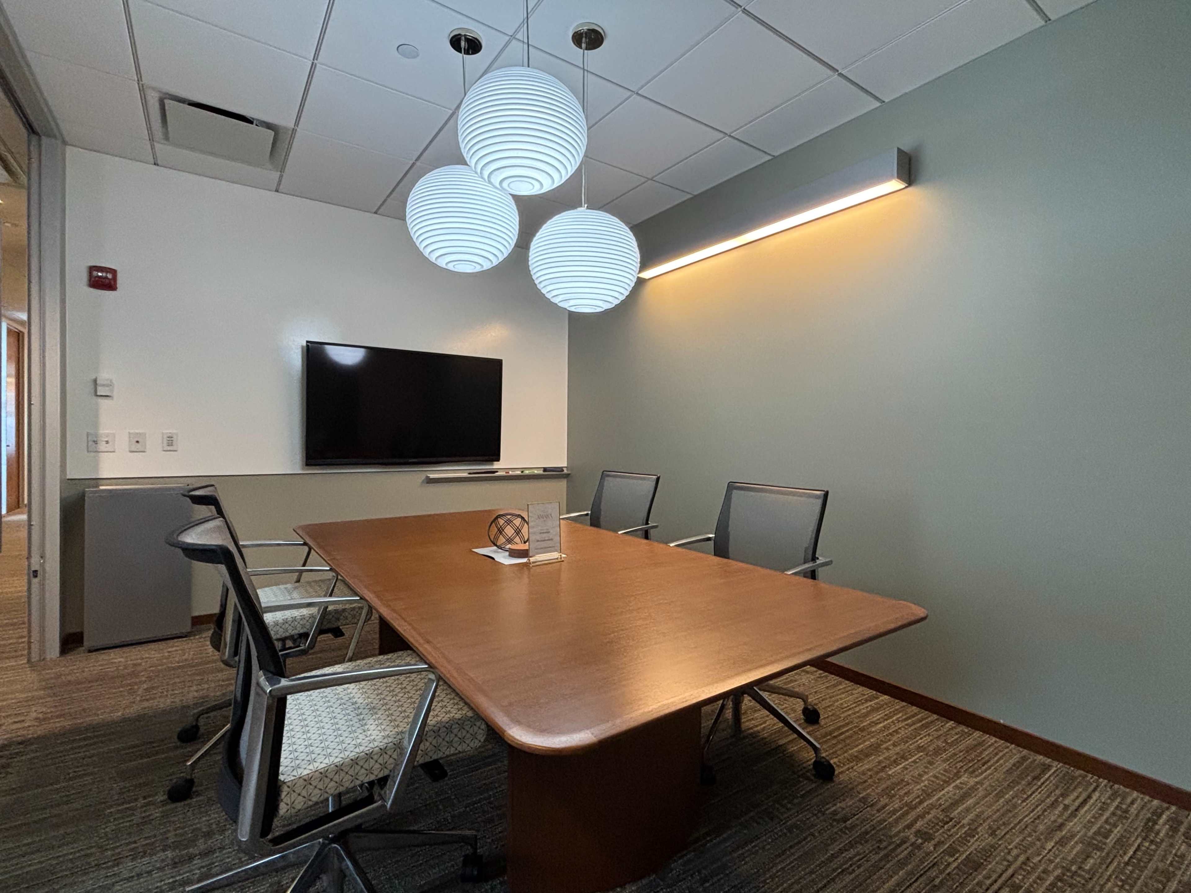 The image shows a small conference room featuring a wooden table, four chairs, a wall-mounted TV, and three pendant lights.