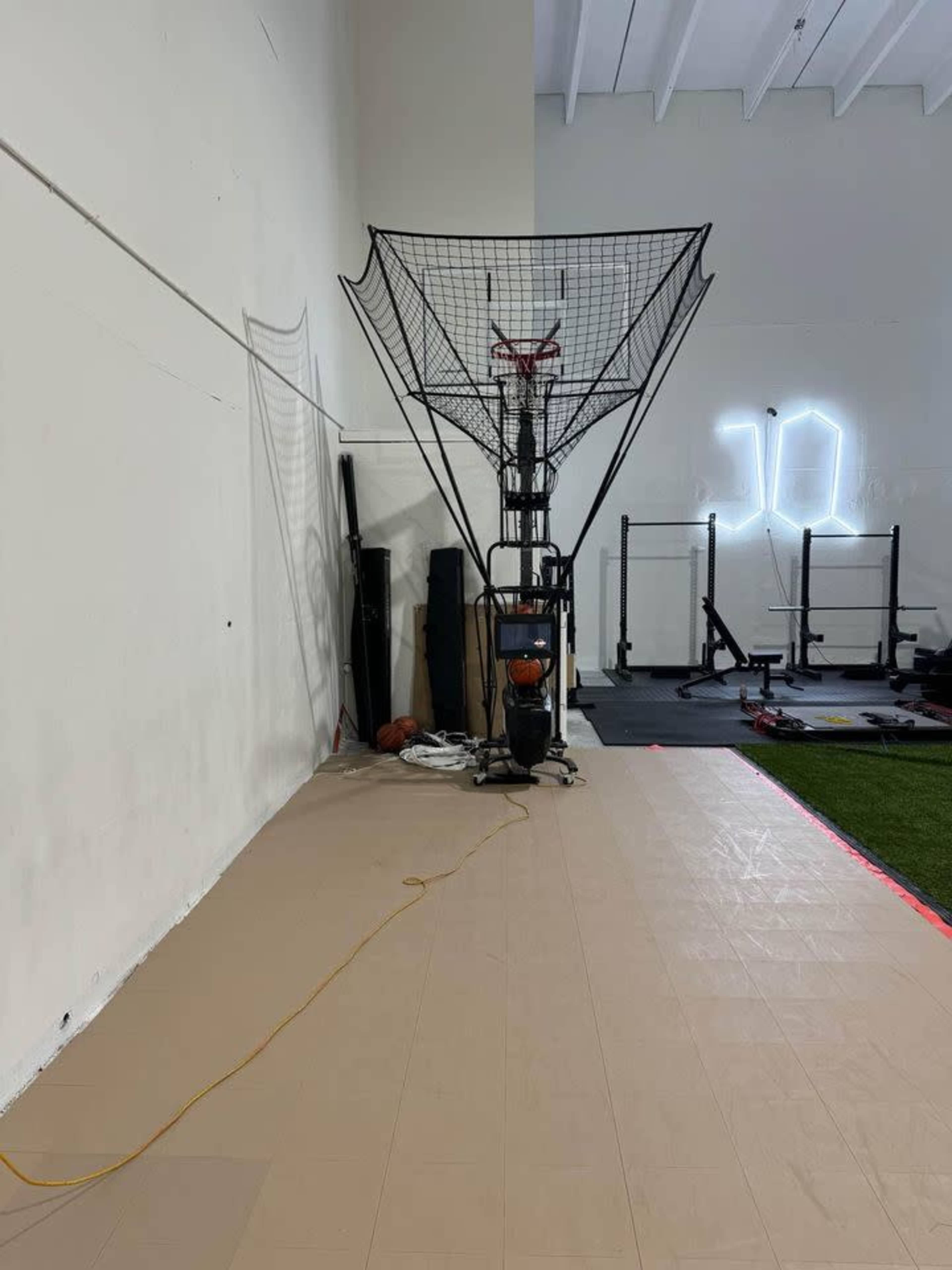 The image shows a basketball shooting machine positioned against a wall in an indoor gym, with various gym equipment visible in the background.