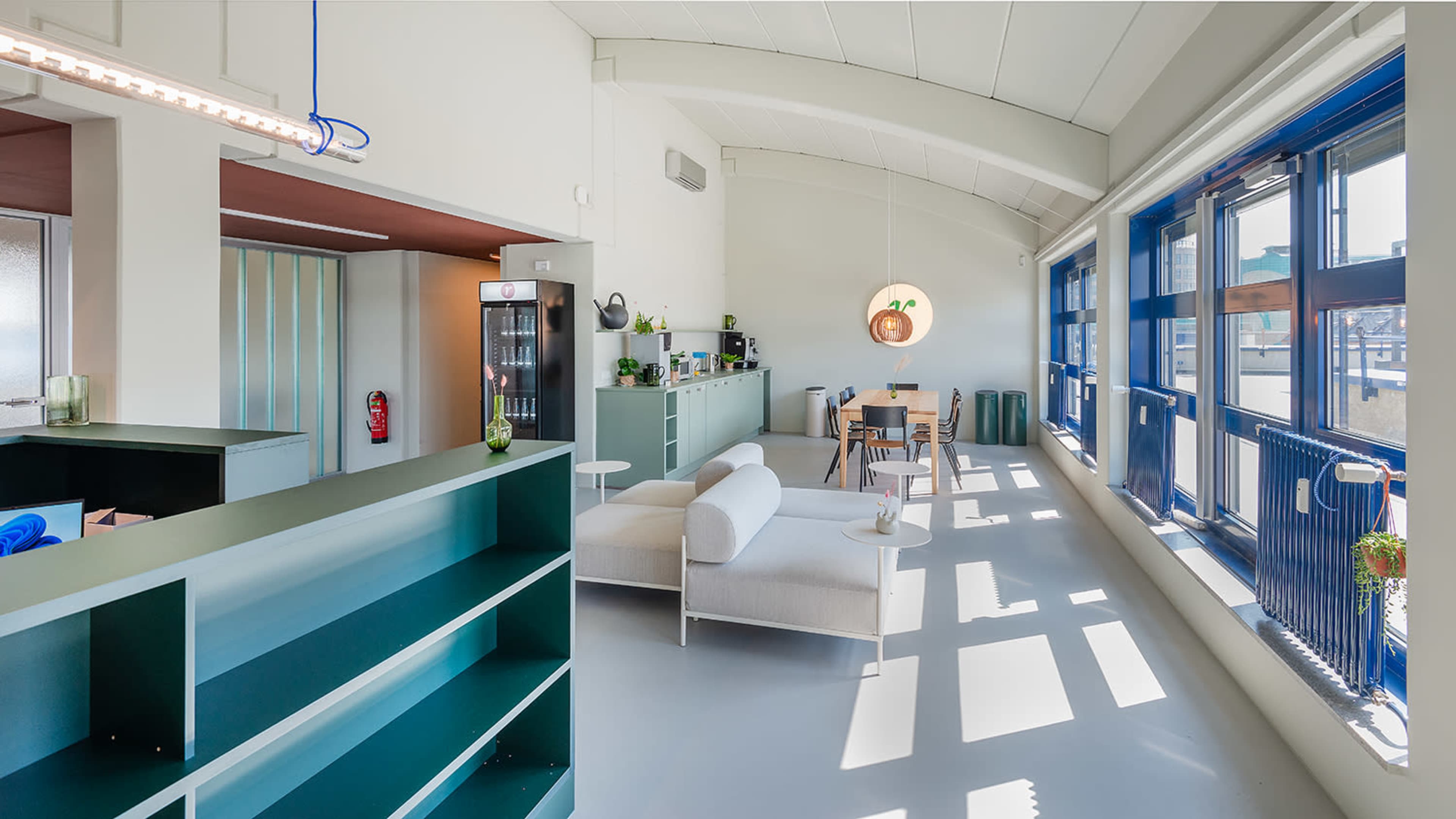 The image shows a modern, bright interior of a living space with a green kitchen area, a dining table, and large windows casting shadows on the floor.