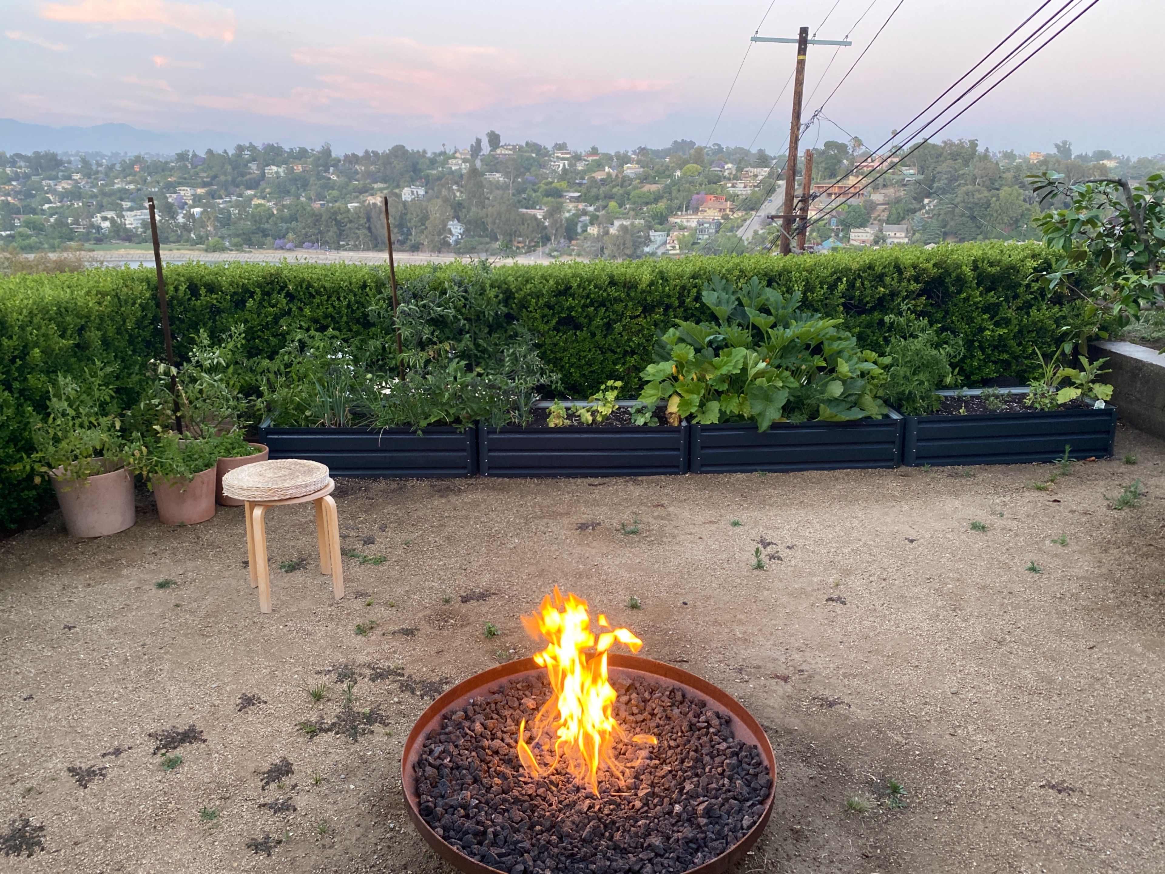 A fire pit with flames is surrounded by black rocks in a garden area with potted plants and a view of hills in the background.