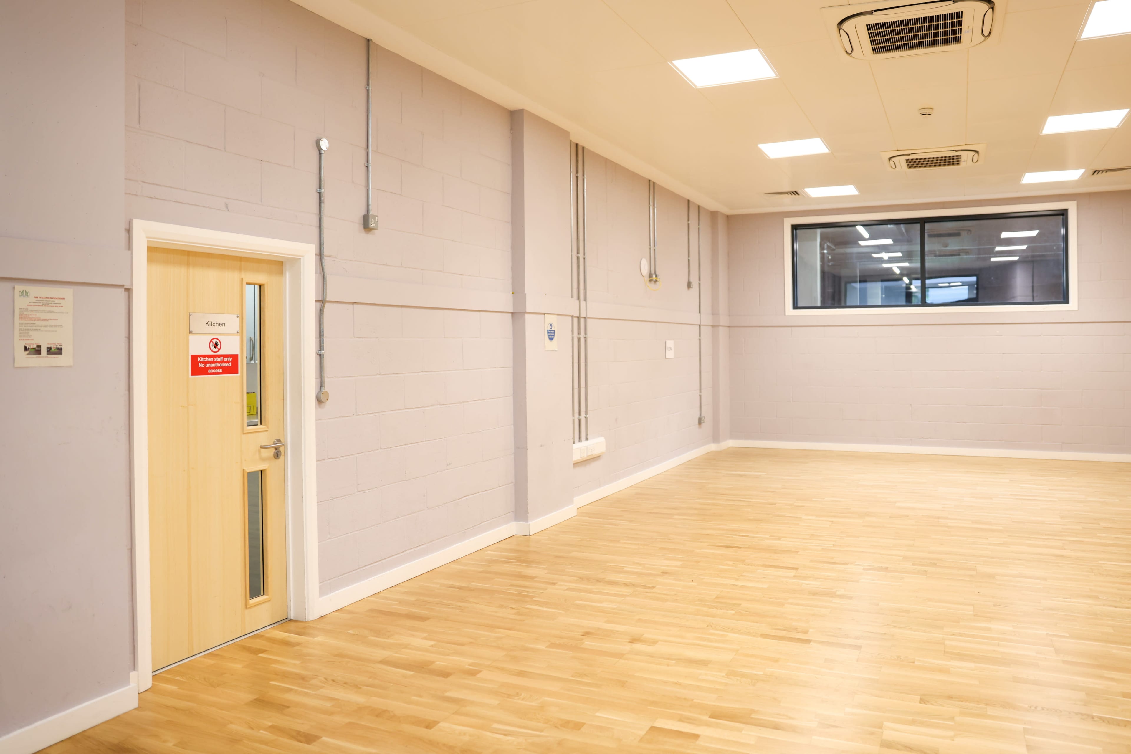 A spacious room with wooden flooring, a single door labeled "Kitchen," and large windows allowing natural light.
