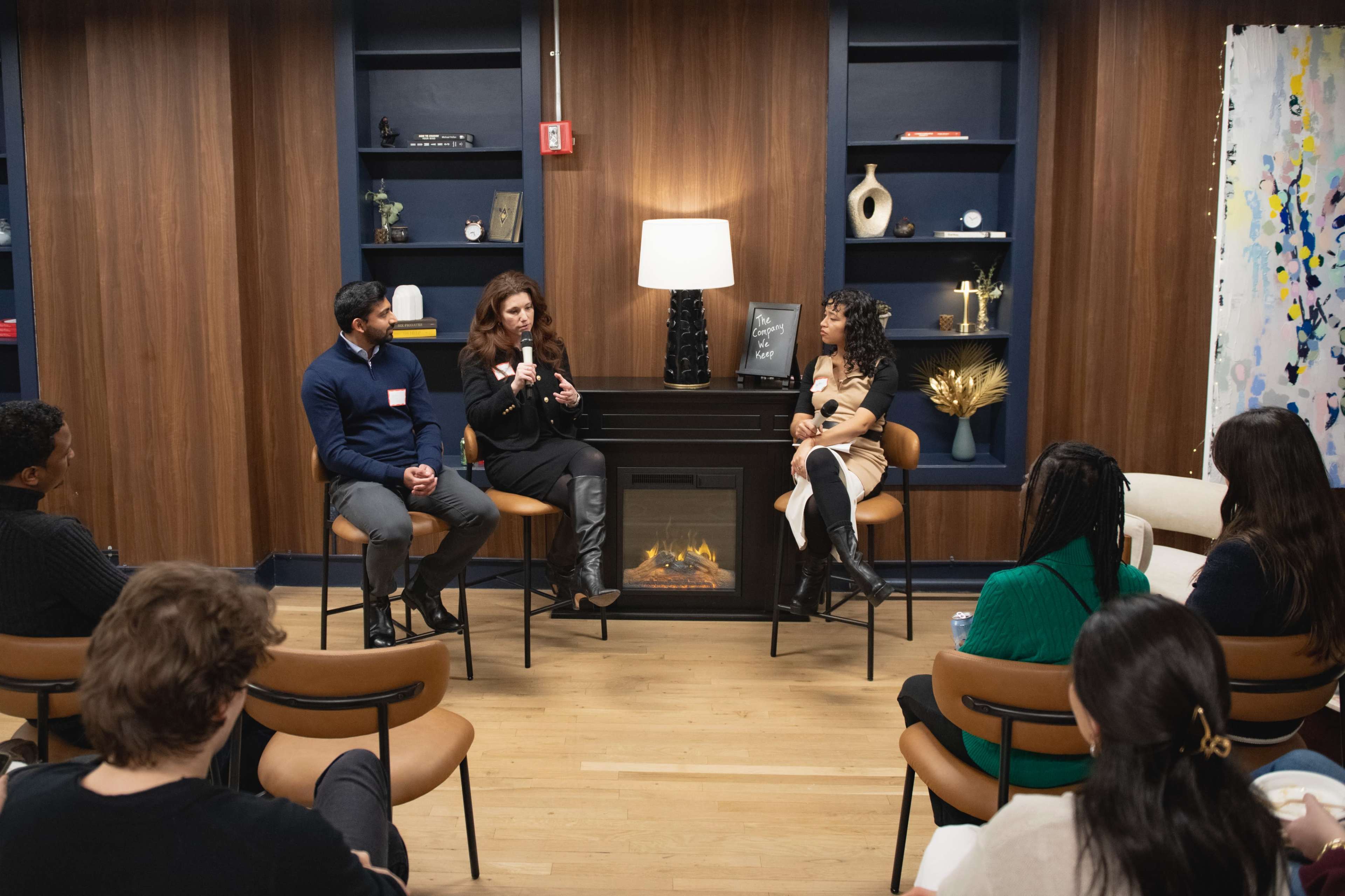 A panel discussion is taking place in a cozy room with wooden walls, featuring two speakers seated on bar stools beside a fireplace, while an audience listens attentively.