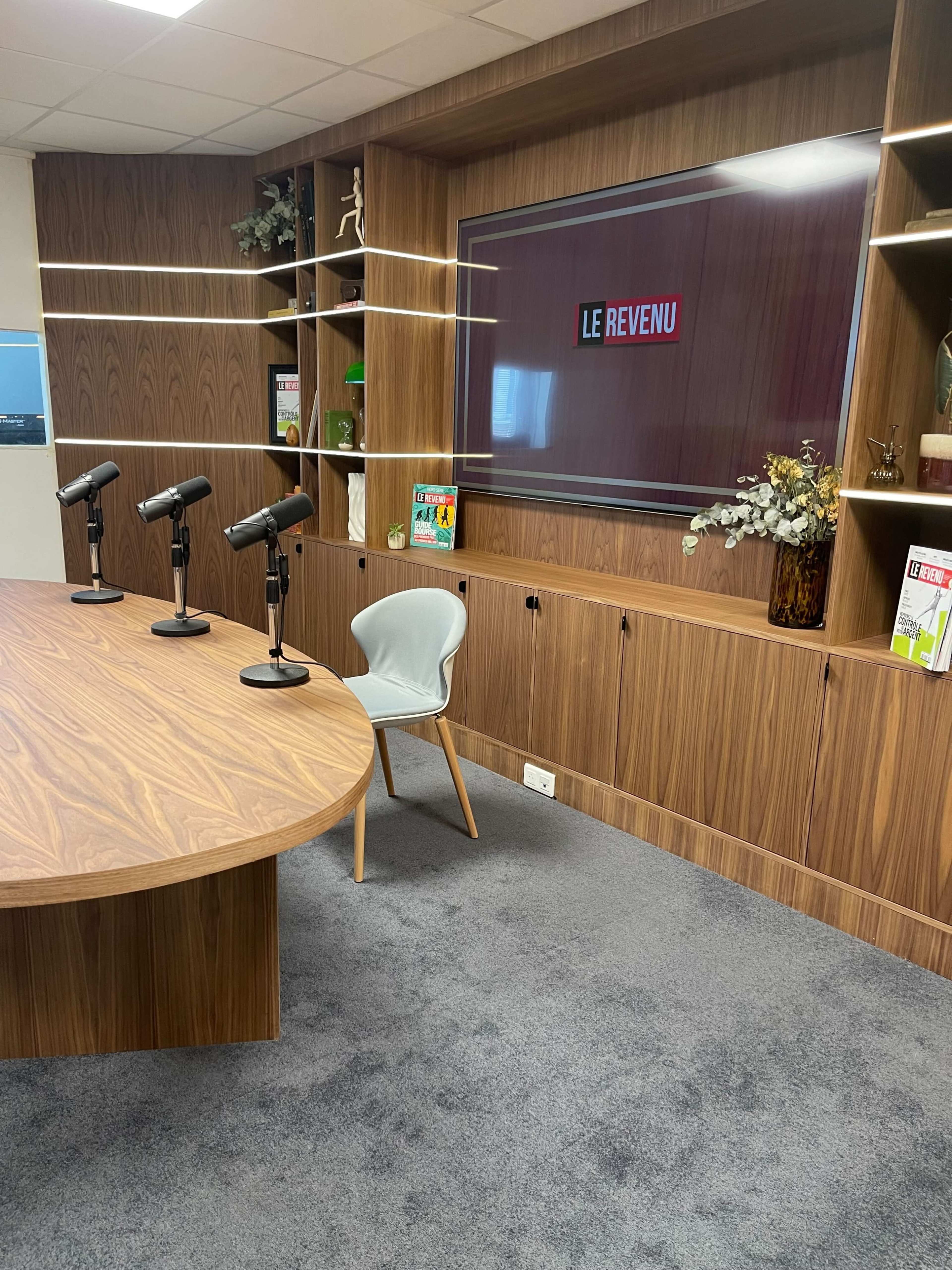 The image shows a modern conference room featuring a large round table, three microphones on the table, a wall-mounted screen displaying "LE REVENU," and wooden paneling with decorative items and bookshelves.