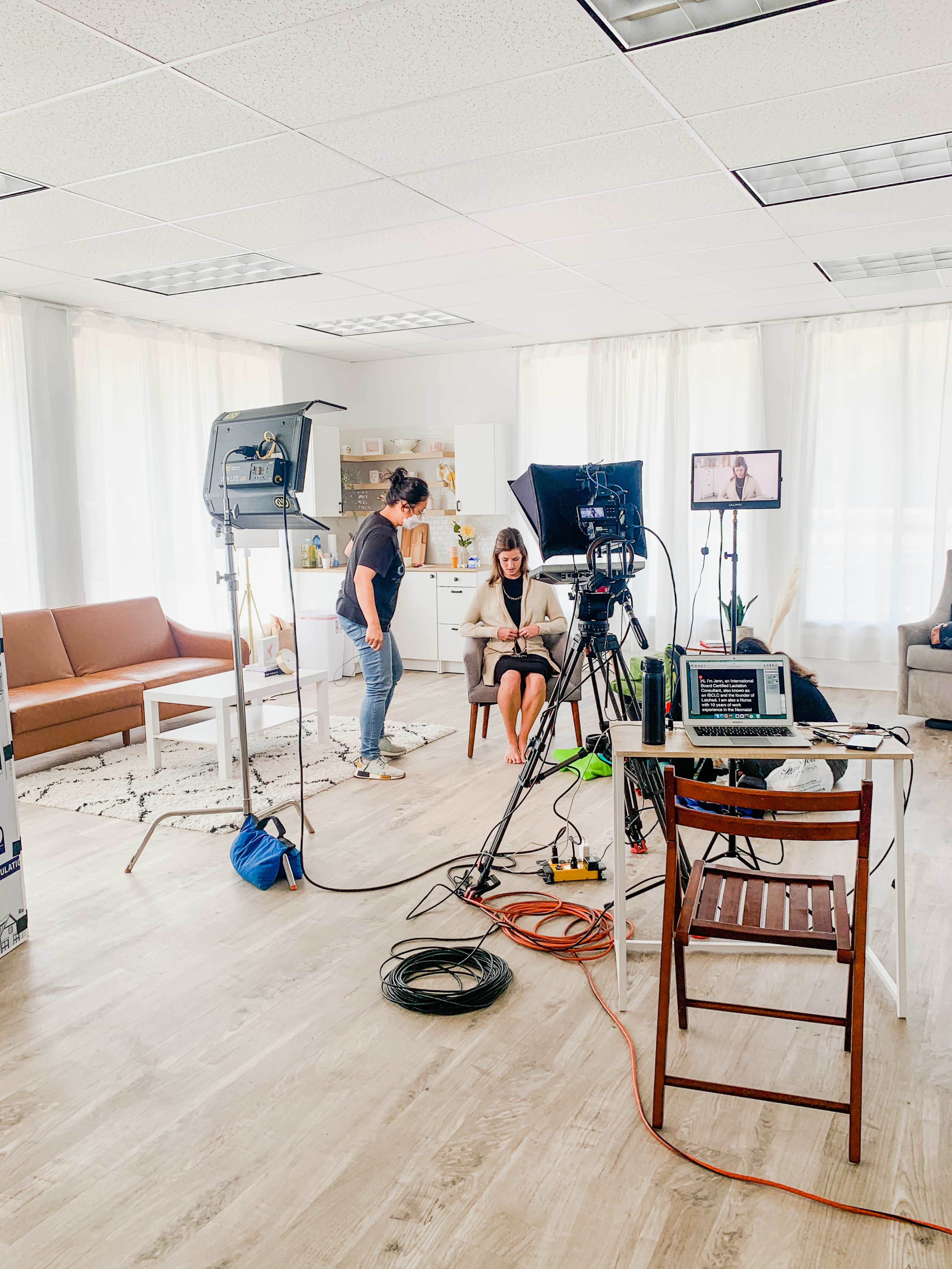 A woman sits on a chair in a bright room set up for filming, with cameras and lights arranged around her.